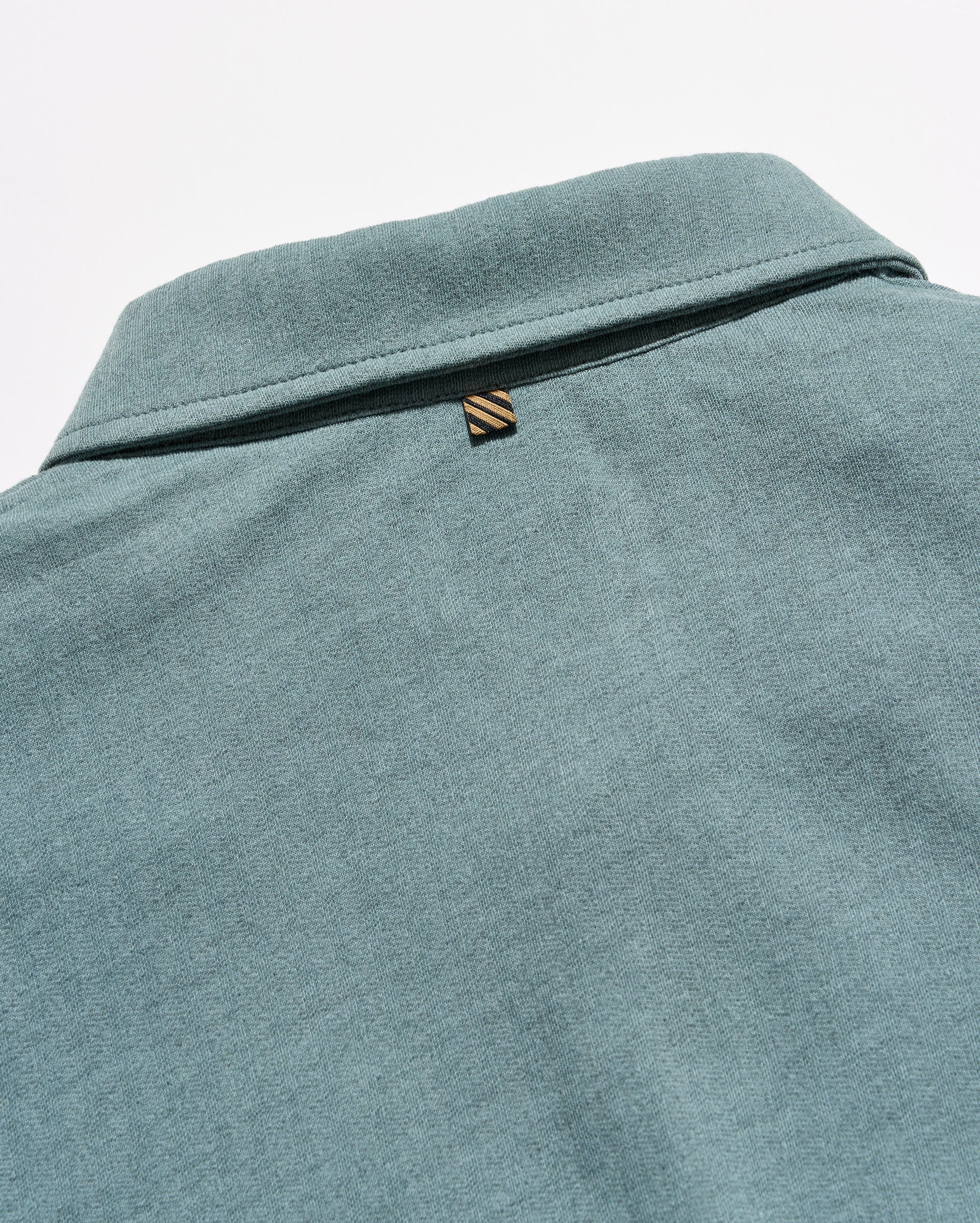Close-up of the back collar of a heathered teal-green polo shirt, featuring a small embroidered striped logo tab.