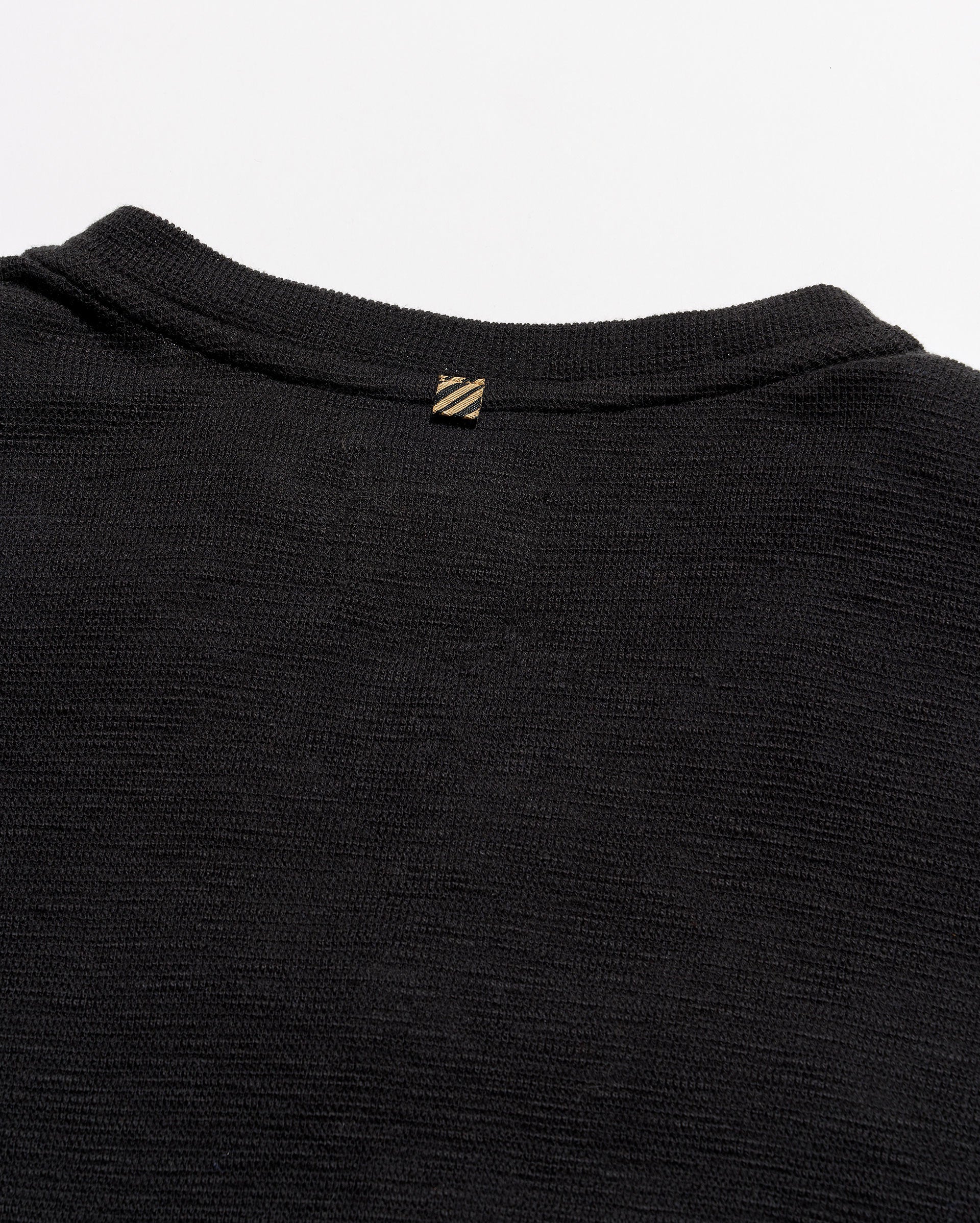Close-up of the back collar of a black ribbed knit sweater, featuring a small gold-tone embroidered logo with diagonal stripes.