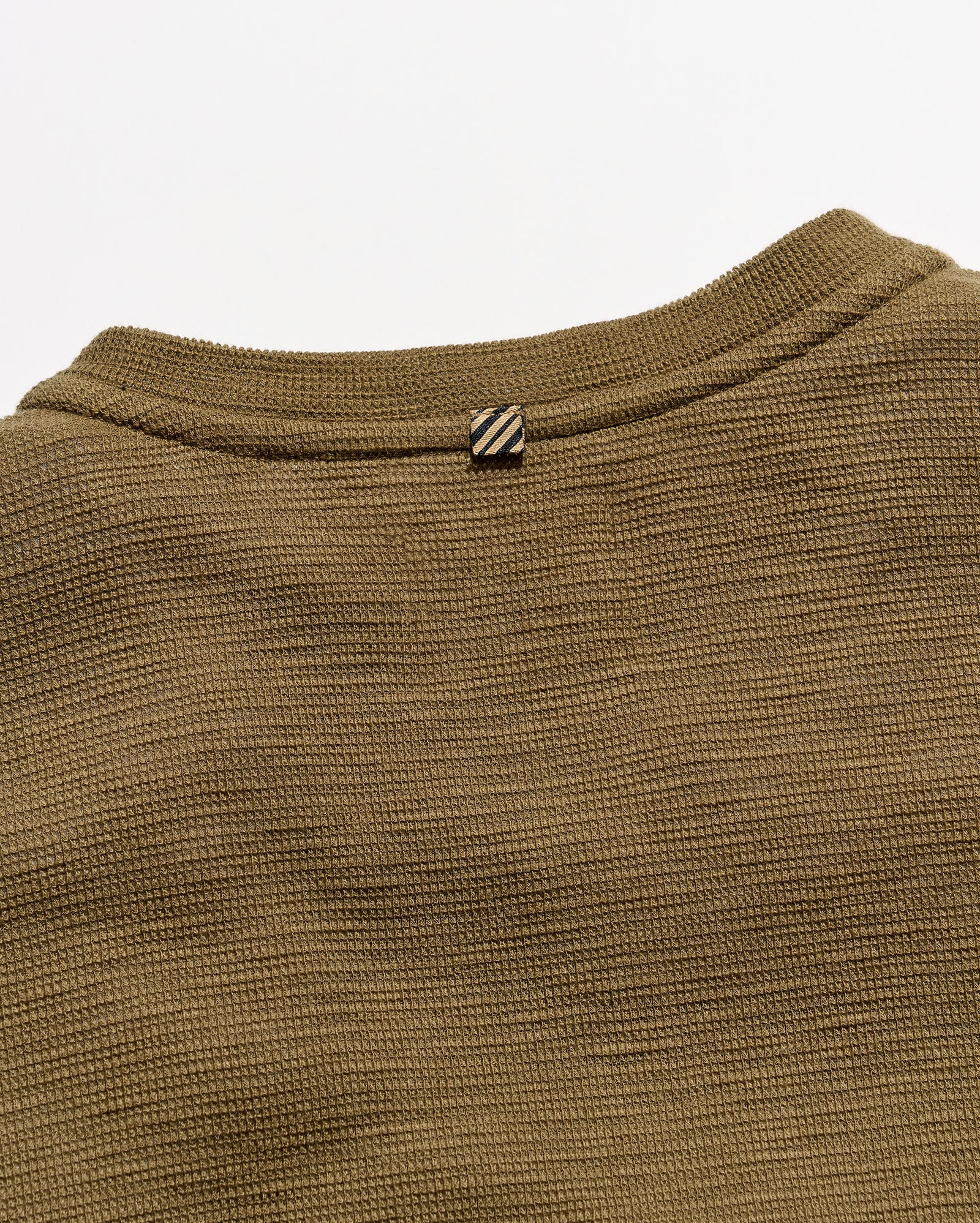 Close-up of a ribbed knit crewneck sweater in olive brown, showing the collar and a small striped detail against a light gray background.