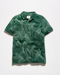 A short-sleeve polo shirt with an abstract green and white pattern, featuring a two-button placket and a classic collar, laid flat against a light gray background.