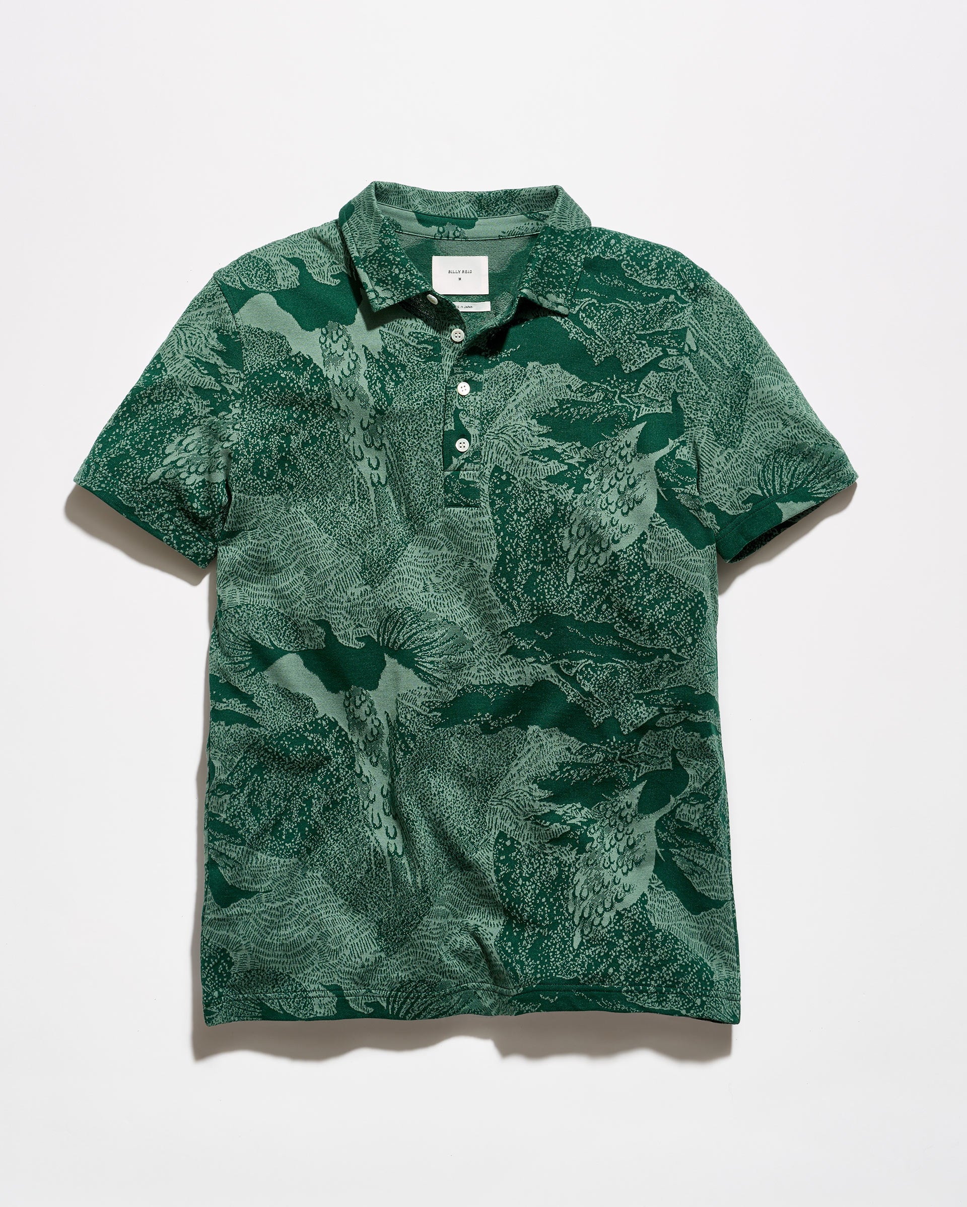 A short-sleeve polo shirt with an abstract green and white pattern, featuring a two-button placket and a classic collar, laid flat against a light gray background.