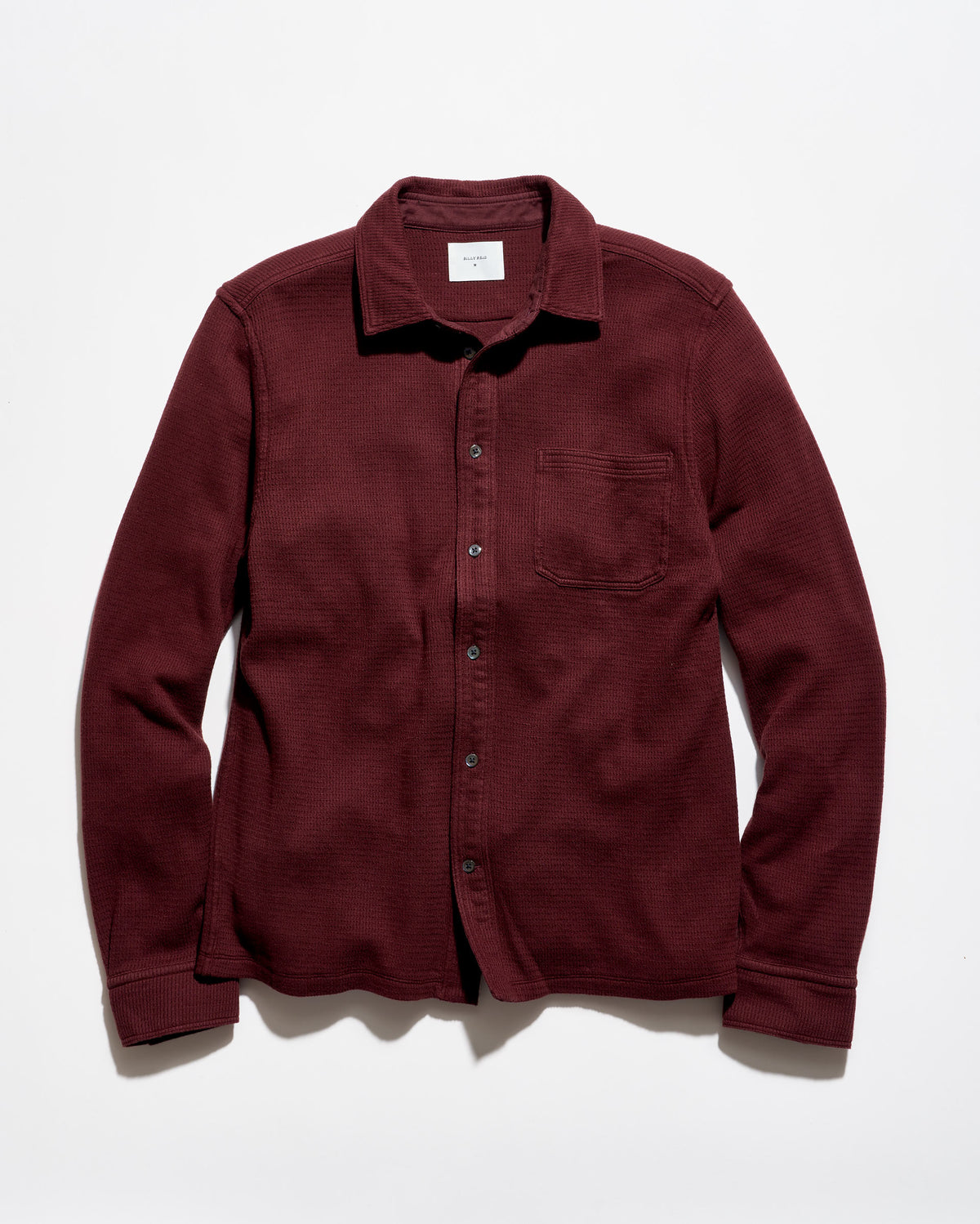 A long-sleeve button-up shirt in a deep burgundy color, laid flat against a light gray background. The shirt features a classic collar, a chest pocket, and dark buttons.