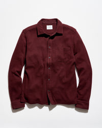 A long-sleeve button-up shirt in a deep burgundy color, laid flat against a light gray background. The shirt features a classic collar, a chest pocket, and dark buttons.
