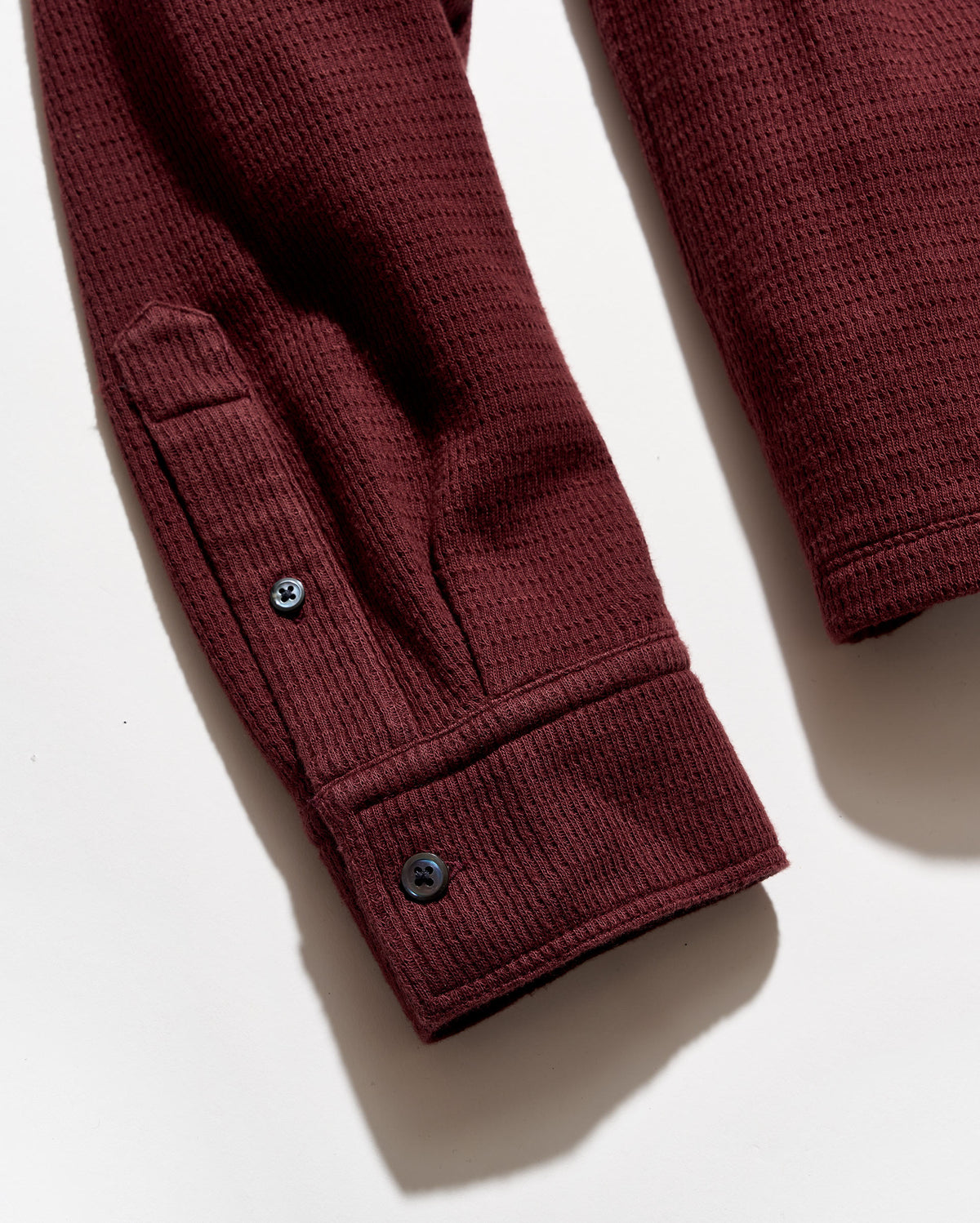 Close-up of a burgundy ladder-knit garment sleeve with a buttoned cuff, photographed on a white background.