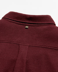 Close-up of a ladder knit button down deep burgundy, showing the collar, a small embroidered logo patch, and the textured fabric.