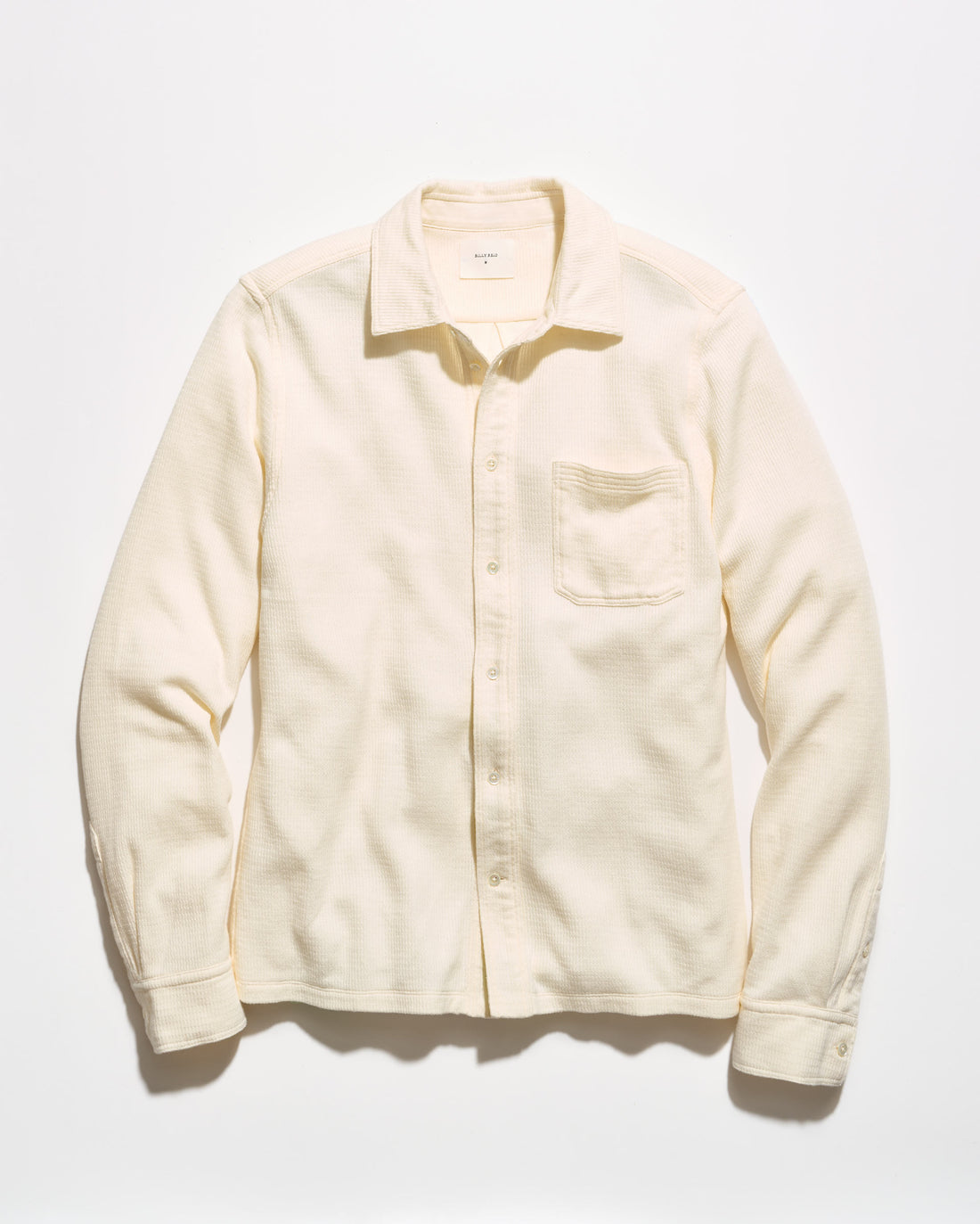A cream-colored long-sleeve button-up shirt with a single chest pocket, laid flat against a light off-white background.