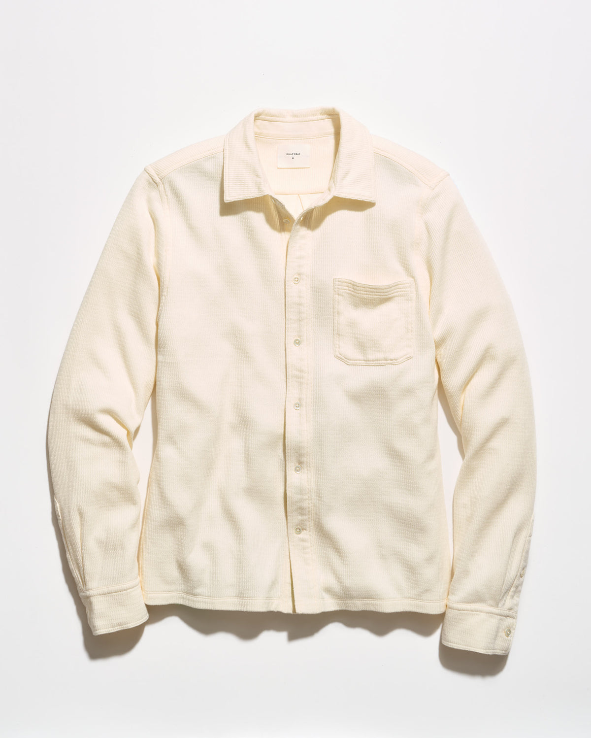 A cream-colored long-sleeve button-up shirt with a single chest pocket, laid flat against a light off-white background.