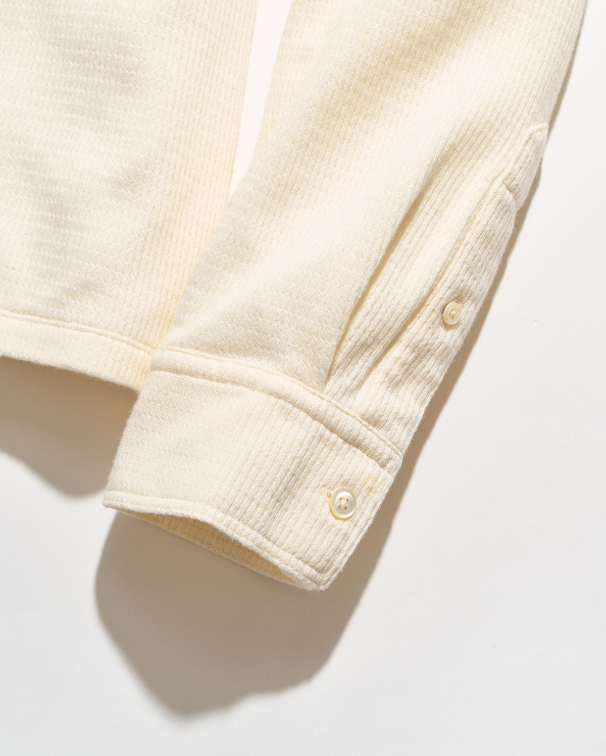 Close-up of a cream-colored ribbed knit garment sleeve with buttoned cuffs, shown on a white background with a soft shadow.