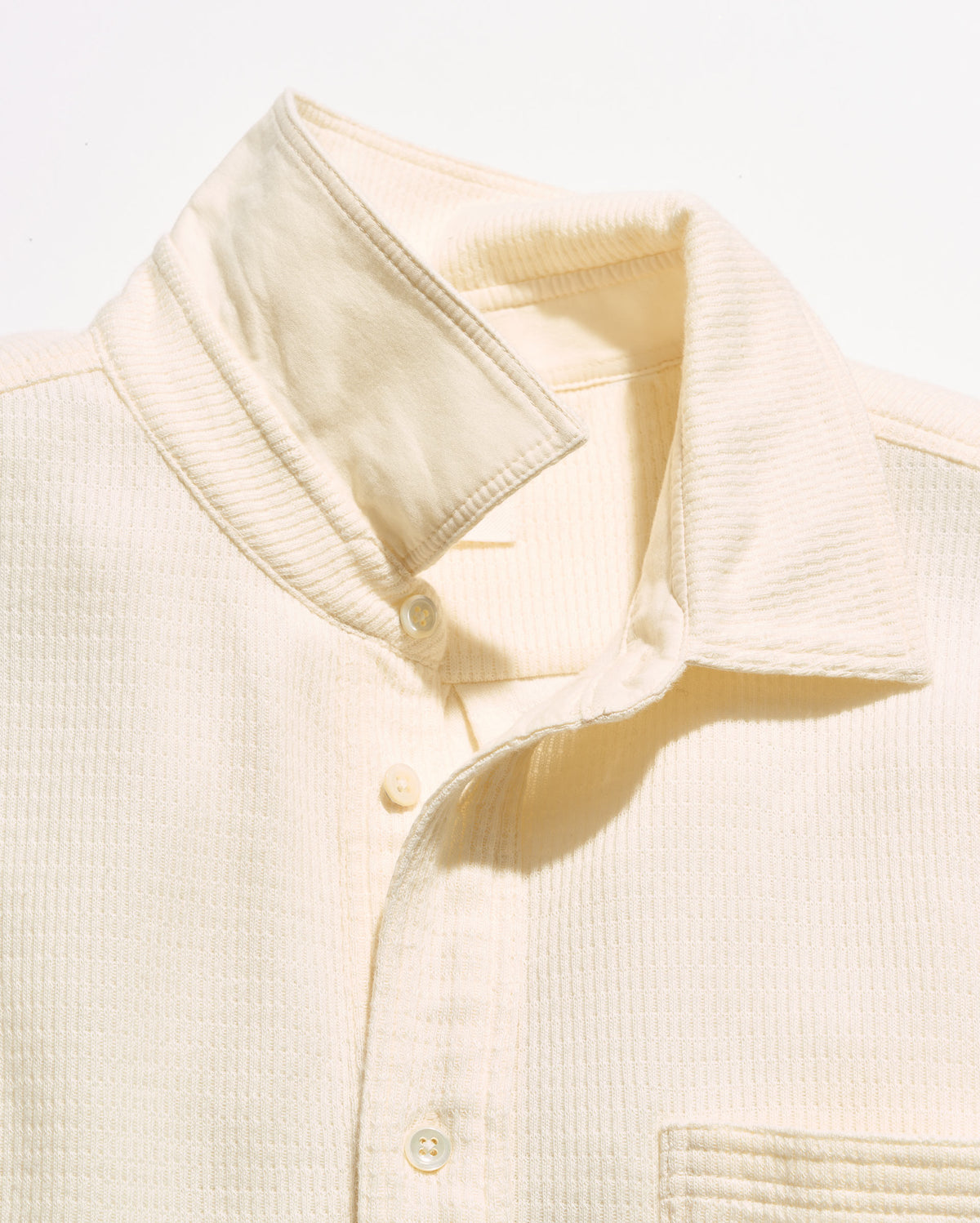 Close-up of a cream-colored textured button-up shirt collar and upper chest, showing the fabric weave and buttons against a plain white background.