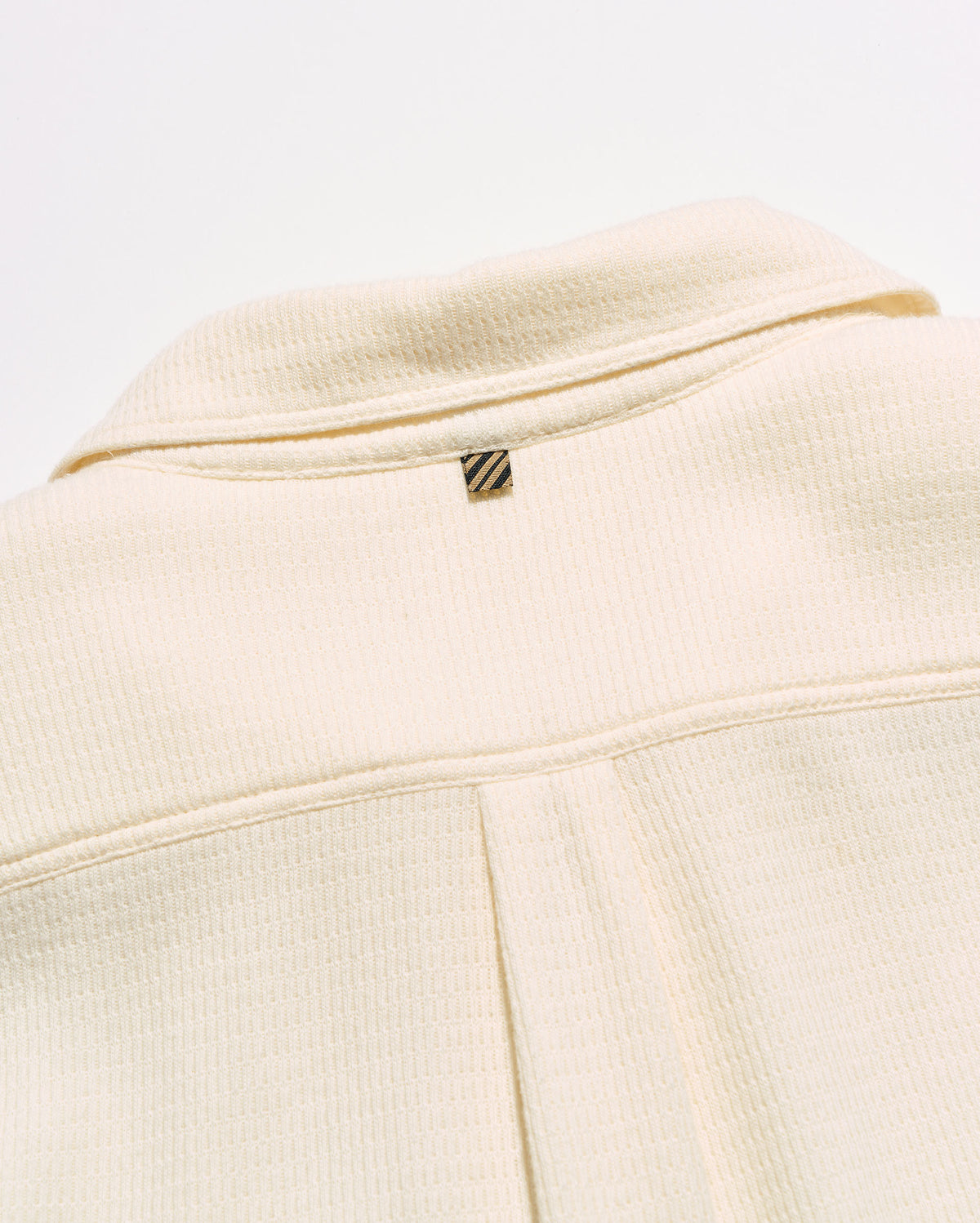 Close-up of a cream-colored textured jacket with a small gold and black striped logo tab near the collar.