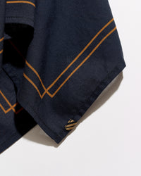 Close-up of a navy blue fabric garment, likely a shirt or jacket sleeve, featuring double diagonal mustard-gold stripes and a small matching embroidered logo near the hem, set against a plain white background.