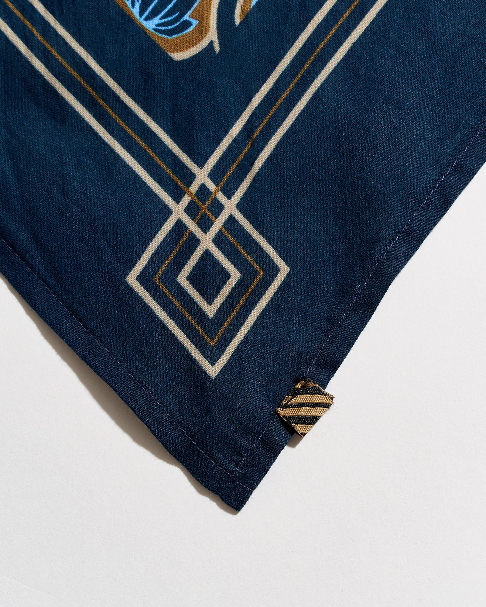 Close-up of a navy blue fabric accessory, likely a scarf or bandana, featuring a geometric diamond pattern in cream and gold, with a small striped fabric tag near the corner.