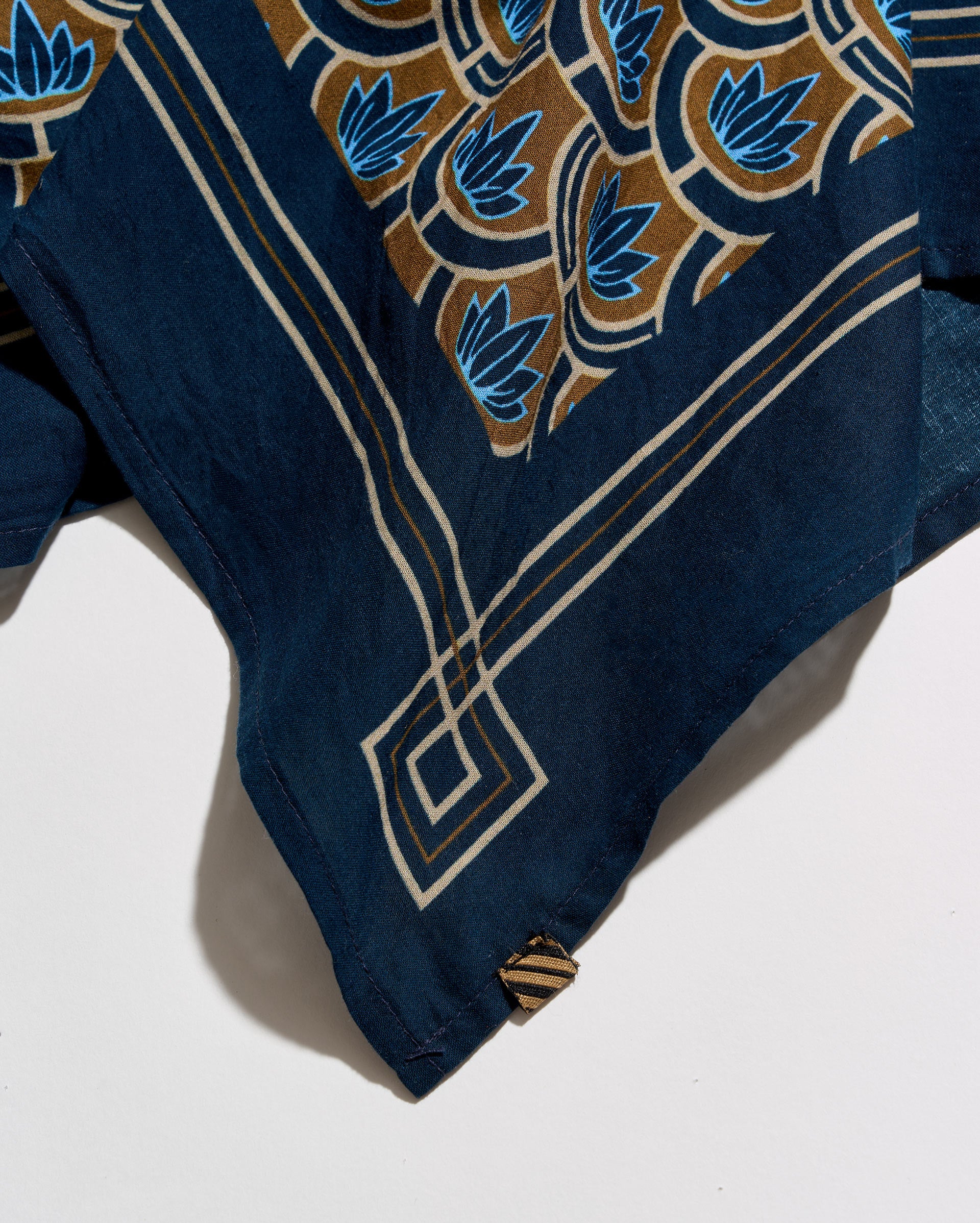 A close-up of a navy blue fabric accessory, likely a scarf or sash, featuring a detailed batik-style pattern in brown, cream, and blue. The fabric is draped diagonally on a clean white background.