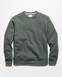 An olive green crewneck sweatshirt with brown leather elbow patches, laid flat against a light gray background.