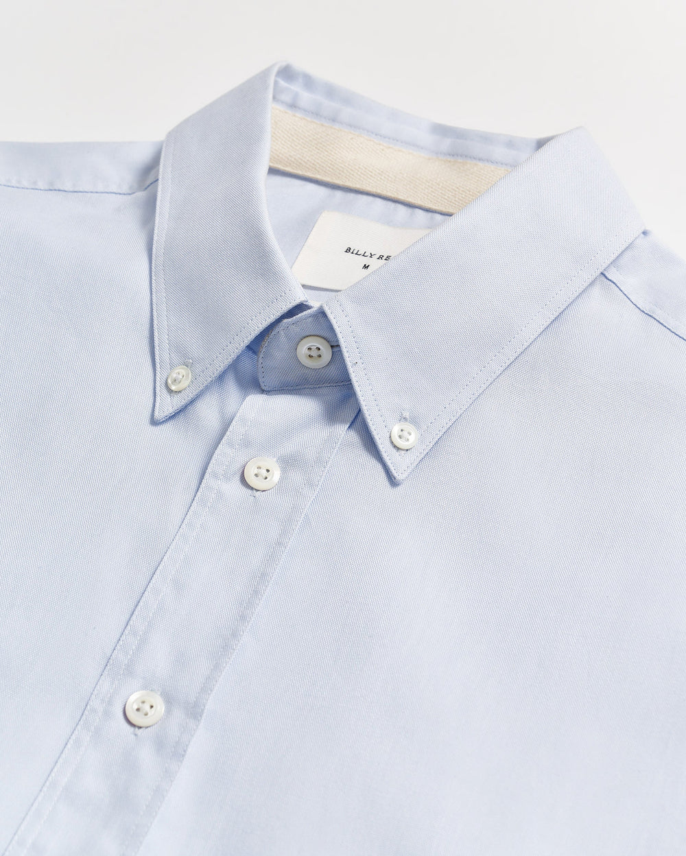 A light blue button-down shirt with white buttons, neatly laid flat on a white surface. The collar is buttoned down, and a white label is partially visible inside the collar.