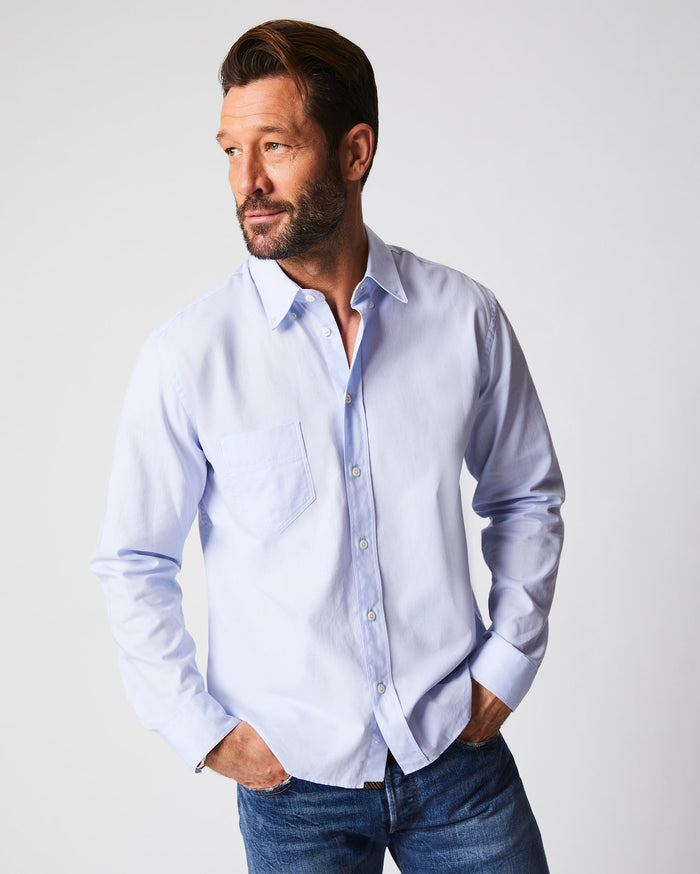 A man with short brown hair and a beard wears a light blue button-up shirt and blue jeans, standing with his hands in his pockets and looking to his left against a plain light background.