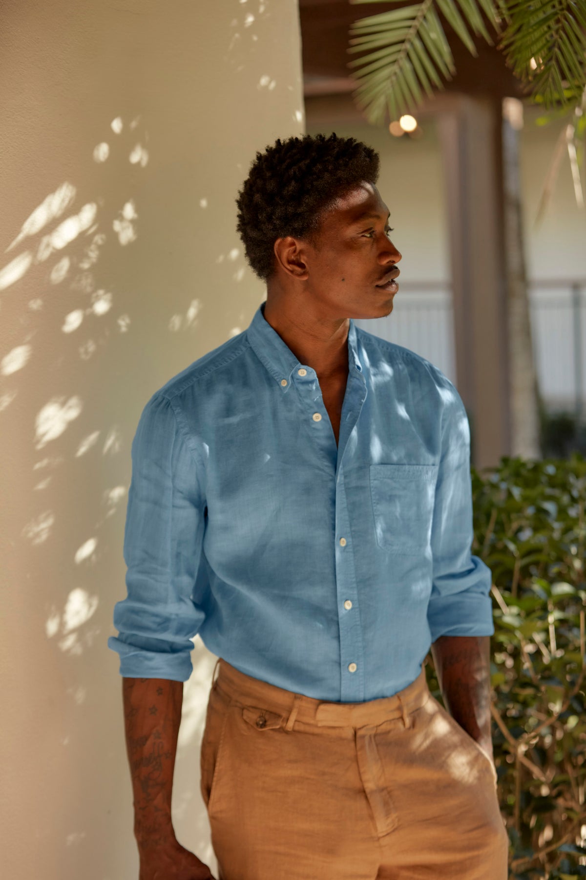 A man stands outdoors beside a light-colored wall, wearing a light blue button-up shirt and tan pants. Sunlight and shadows from nearby leaves fall across him as he looks to the side thoughtfully.