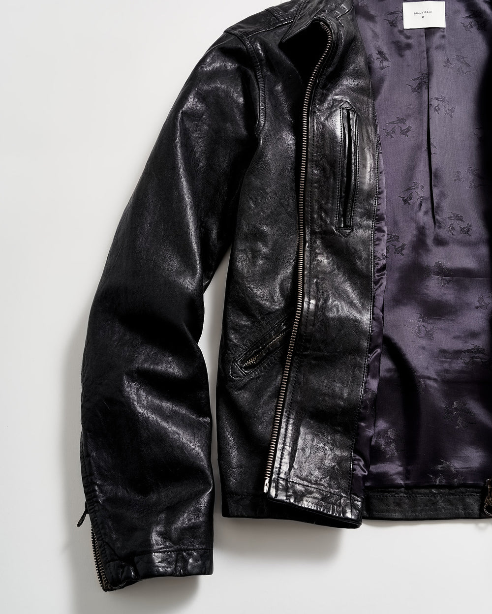 A black leather jacket with a shiny finish is laid flat on a white surface. The jacket is partially unzipped, revealing a purple satin-like inner lining.