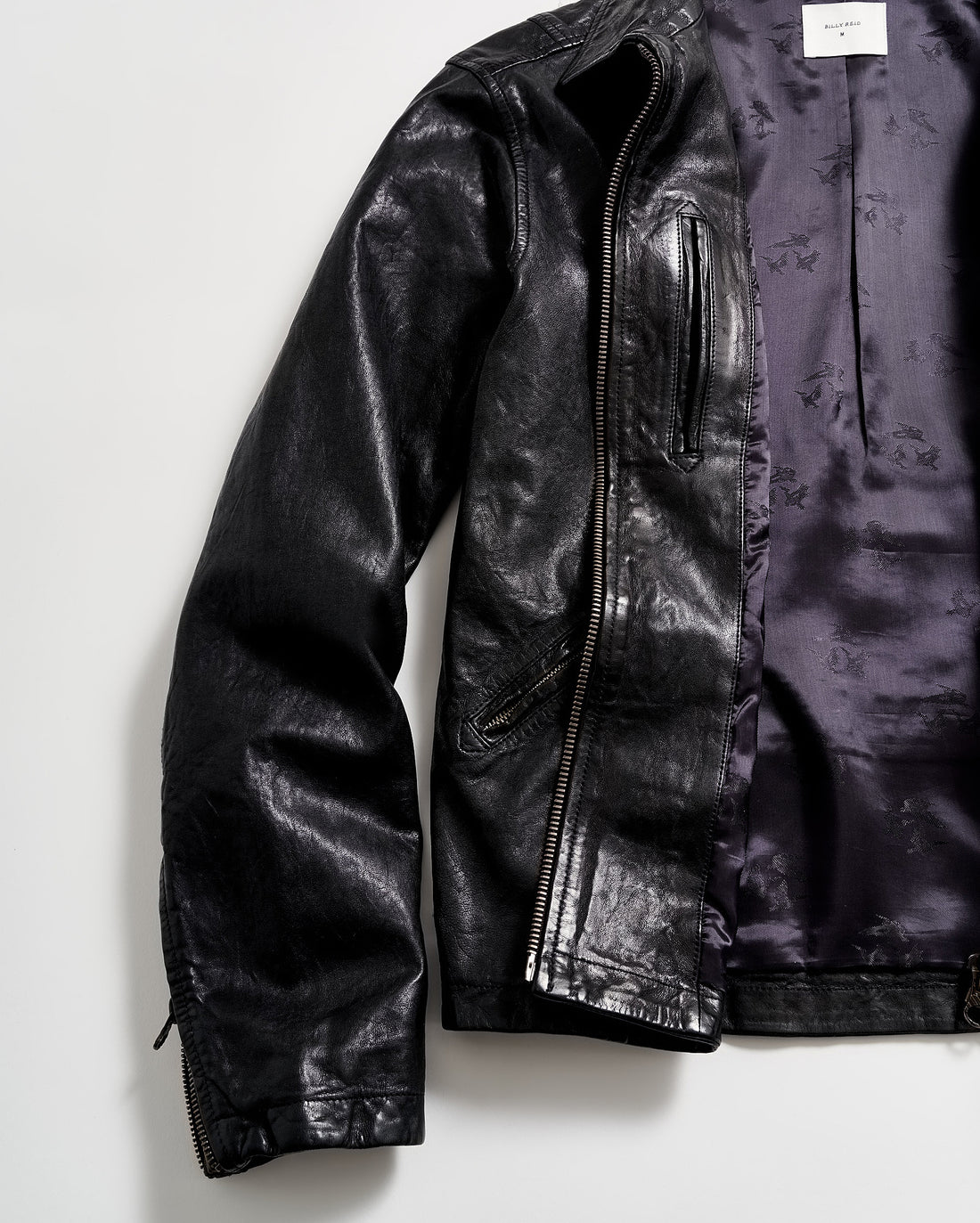 A black leather jacket with a shiny finish is laid flat on a white surface. The jacket is partially unzipped, revealing a purple satin-like inner lining.
