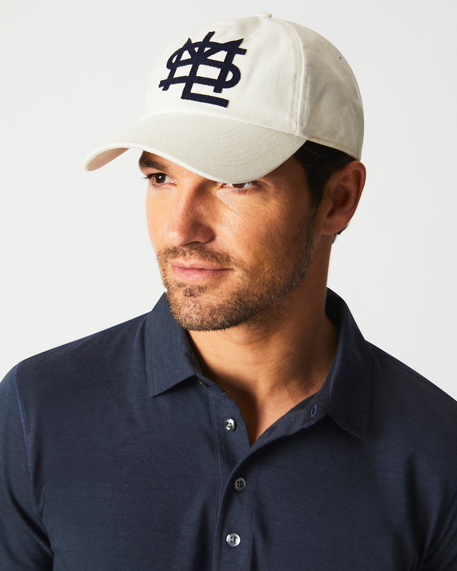 A man wearing a cream-colored baseball cap with a navy blue embroidered logo and a navy blue collared shirt looks slightly to the side against a plain background.