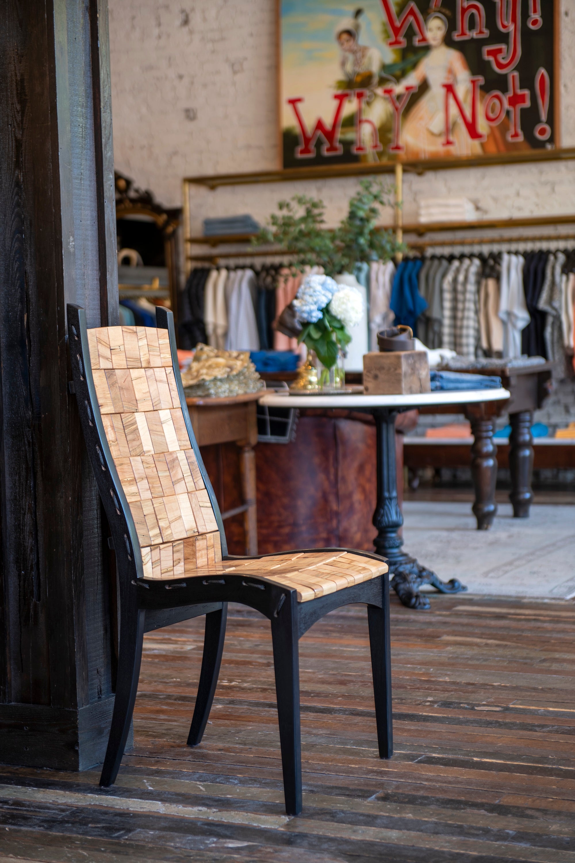 A rustic wooden dining chair with a black metal frame and woven natural-fiber seat and back, positioned on a dark wooden floor in front of a shop interior with clothing racks and a vintage sign.