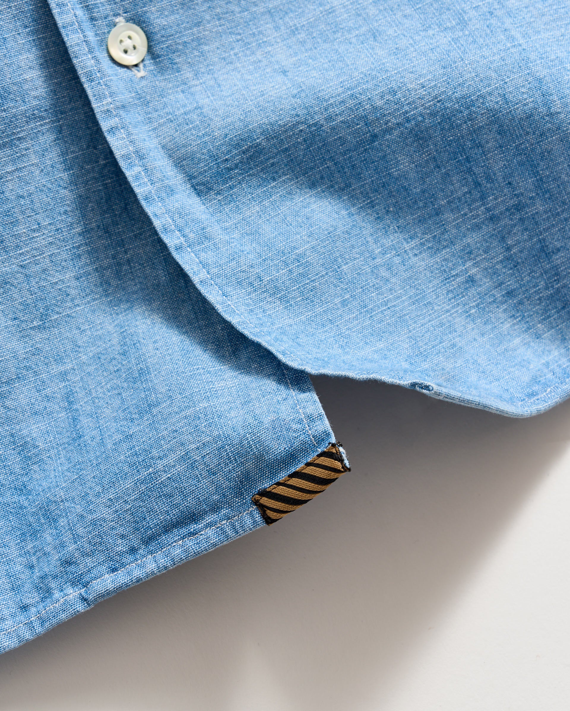 A close-up of the bottom hem of a light blue button-up shirt, showing a white button and a small brown and black striped fabric tag.
