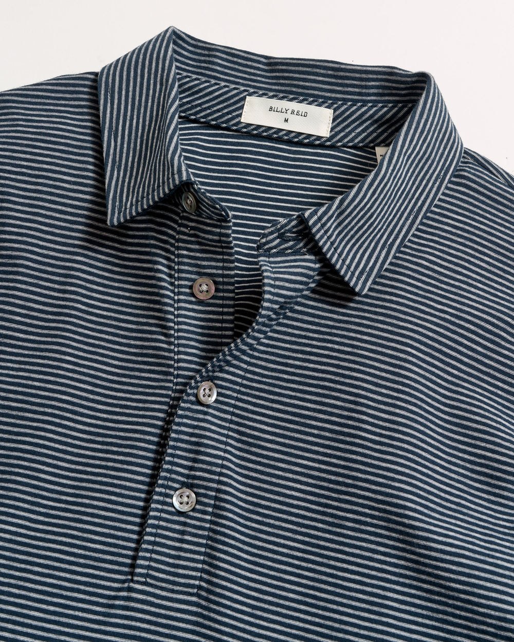A navy blue polo shirt with thin white horizontal stripes, a collar, and a three-button placket, laid flat on a white surface. The label inside the collar reads BILLY REID.