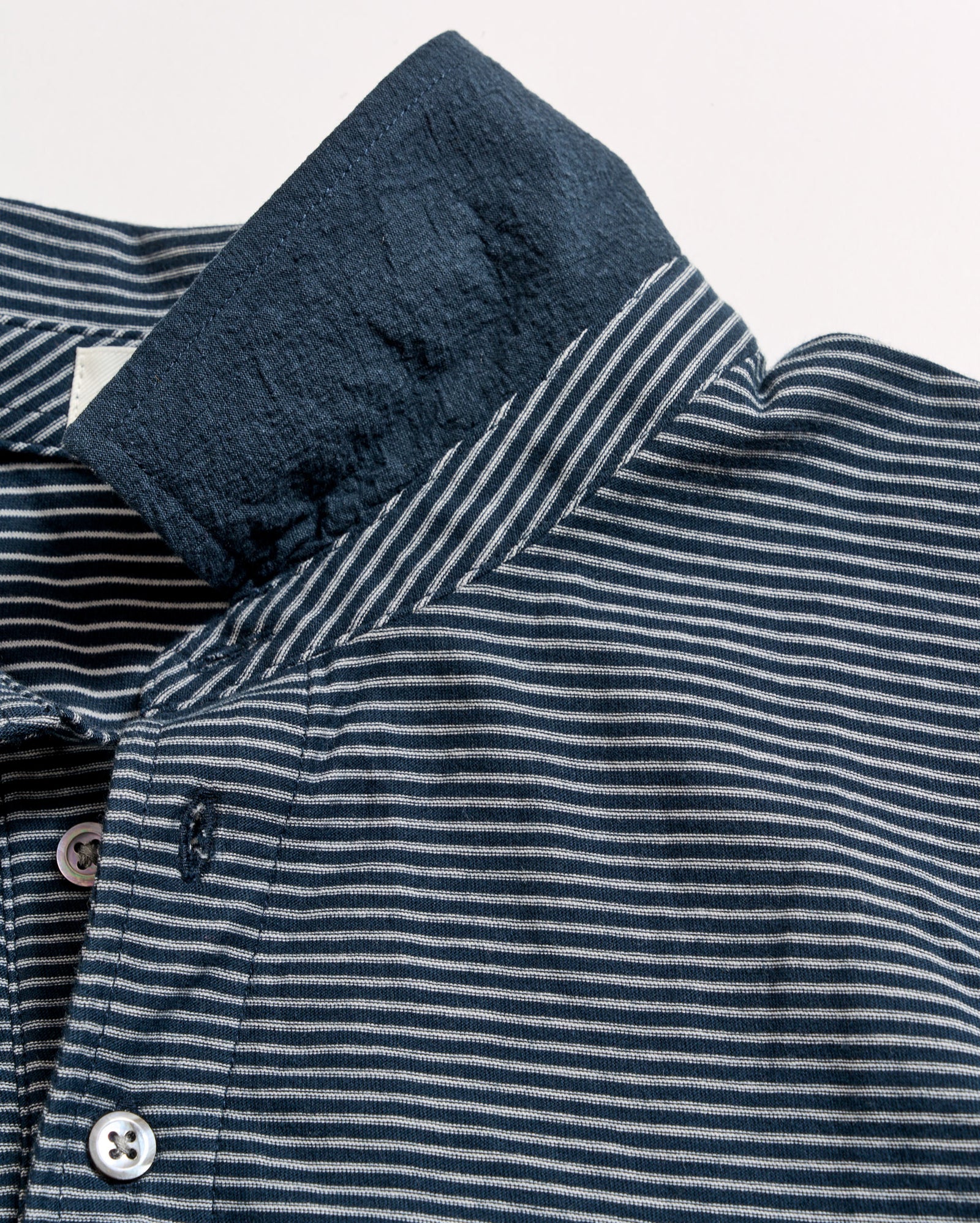 Close-up of a blue and white striped shirt with the collar flipped up, revealing a contrasting solid navy underside. The shirt features white buttons and fine horizontal stripes.