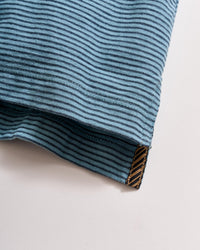 A close-up of a blue fabric with thin black horizontal stripes, showing a neatly stitched hem and a small brown tag at the edge with a diagonal pattern.