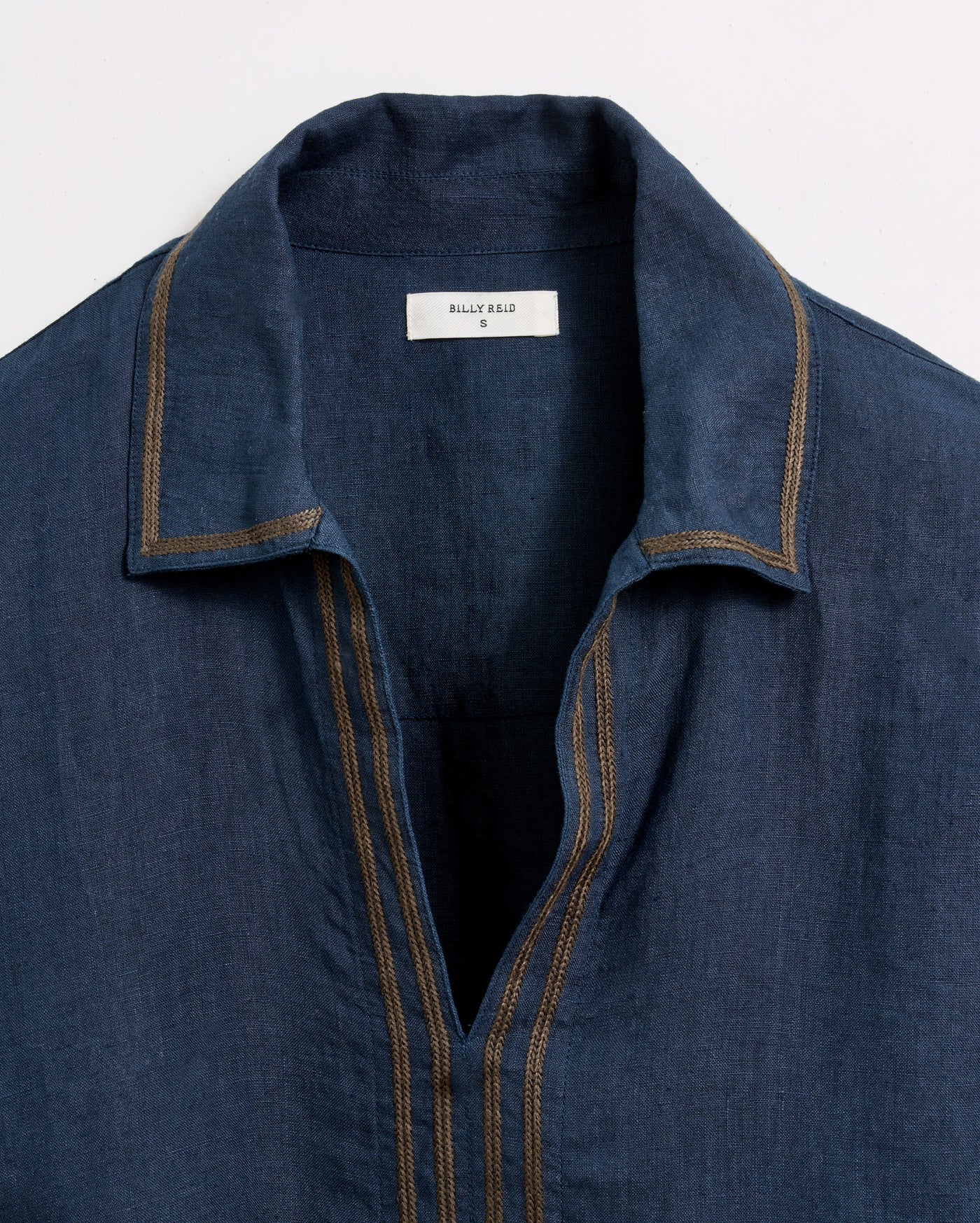 A close-up of a blue collared shirt with tan stitching and an open neckline. The label inside the collar reads “Billy Reid S.” The shirt is laid flat against a white background.