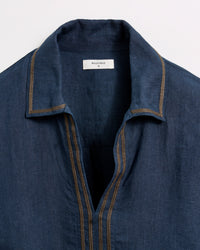 A close-up of a blue collared shirt with tan stitching and an open neckline. The label inside the collar reads “Billy Reid S.” The shirt is laid flat against a white background.