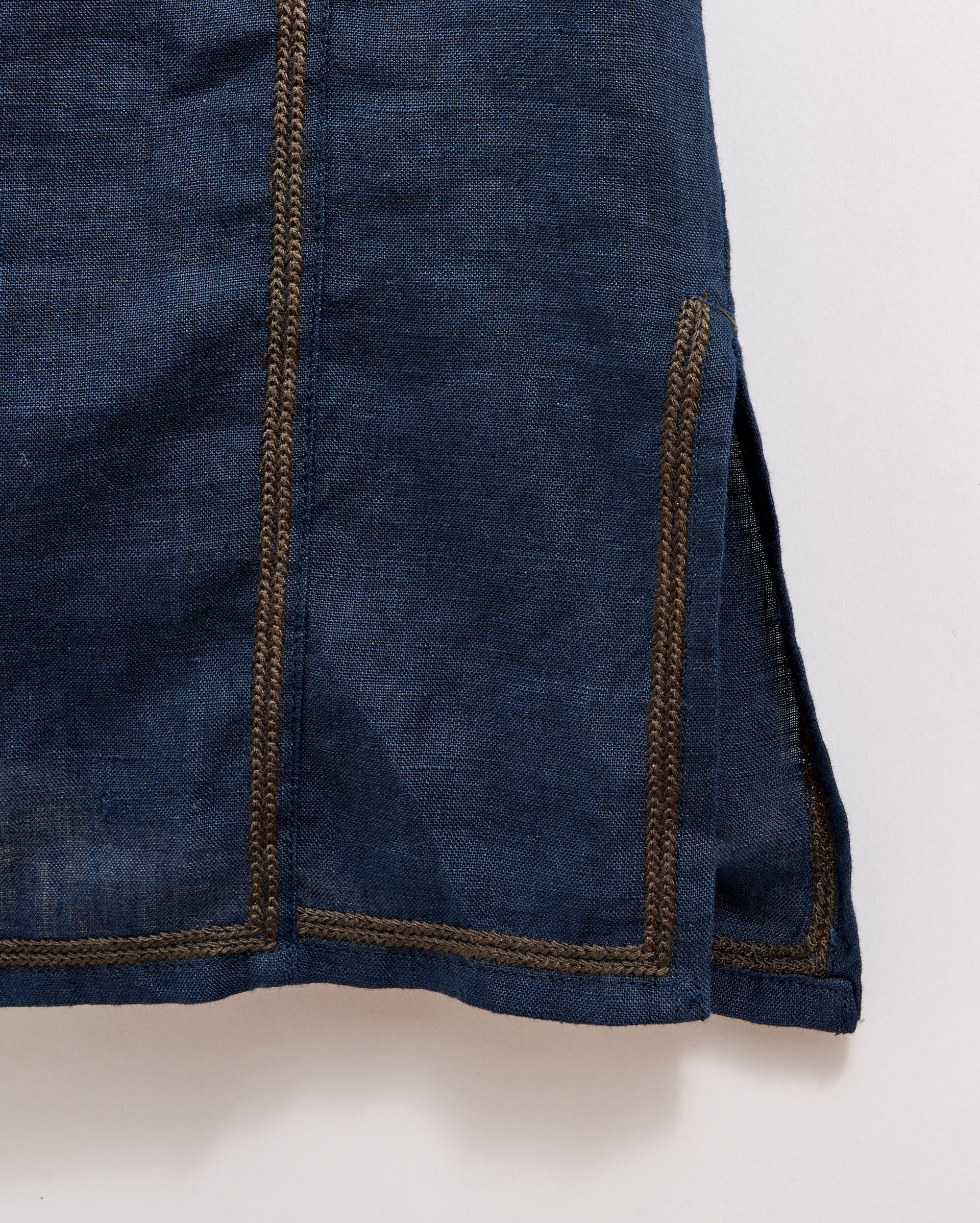 A close-up of a dark blue fabric garment with brown stitched seams and a small side slit near the hemline, photographed against a light background.