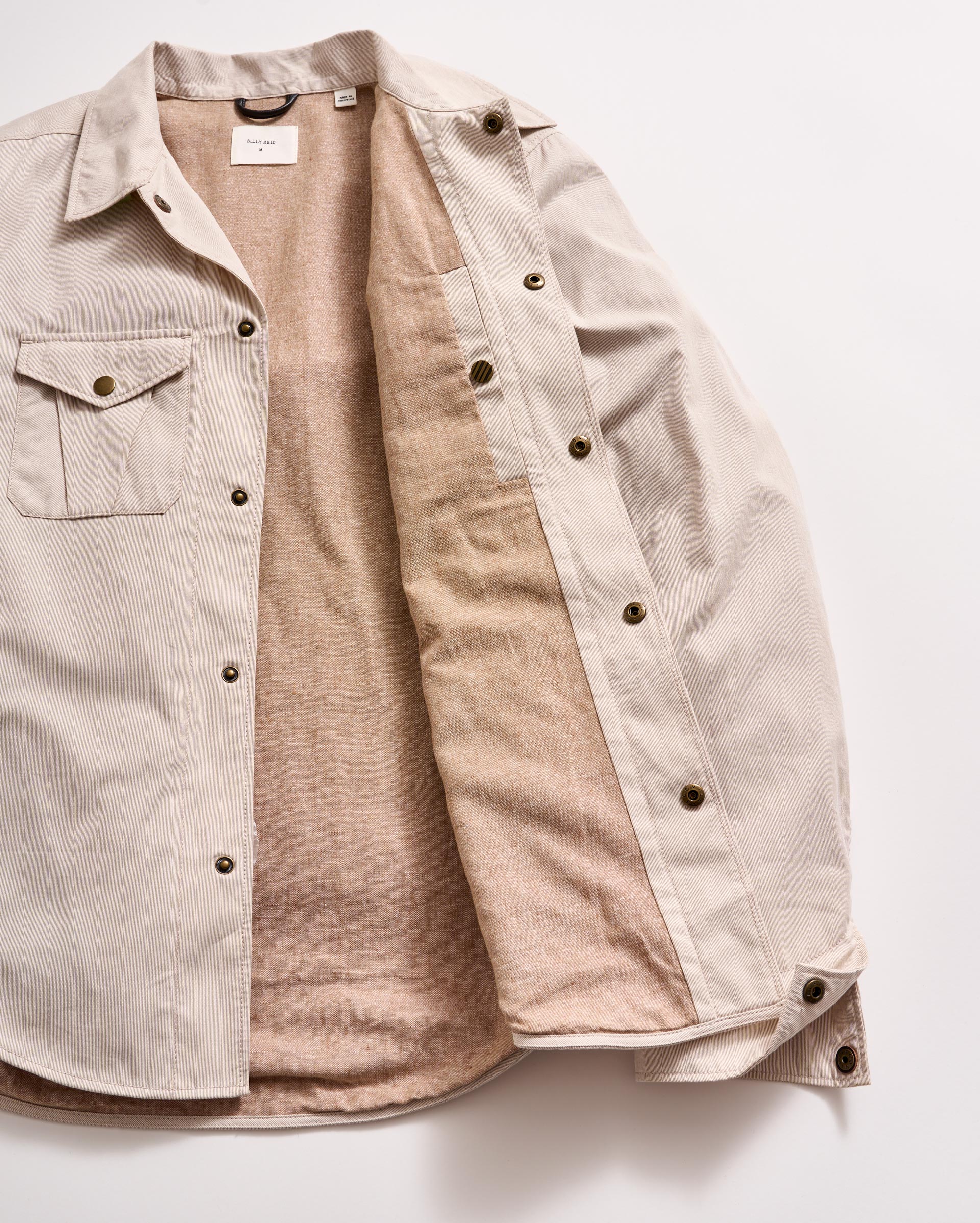 A light beige button-up shirt jacket with brown buttons, one chest pocket, and a soft tan lining, displayed open on a white background.