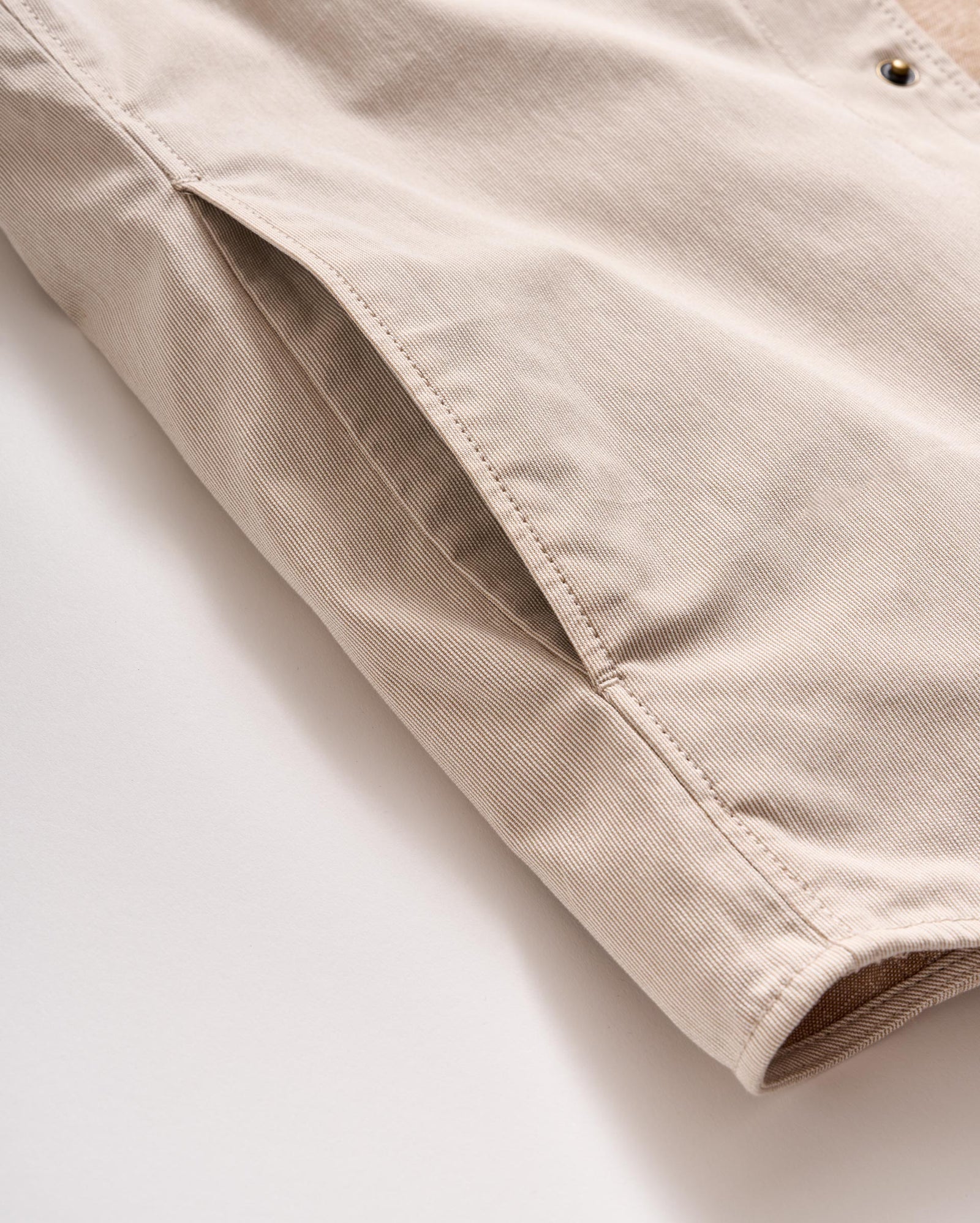 A close-up of a beige fabric jacket, focusing on a side pocket and part of the sleeve, with visible stitching and metal snap button detail.