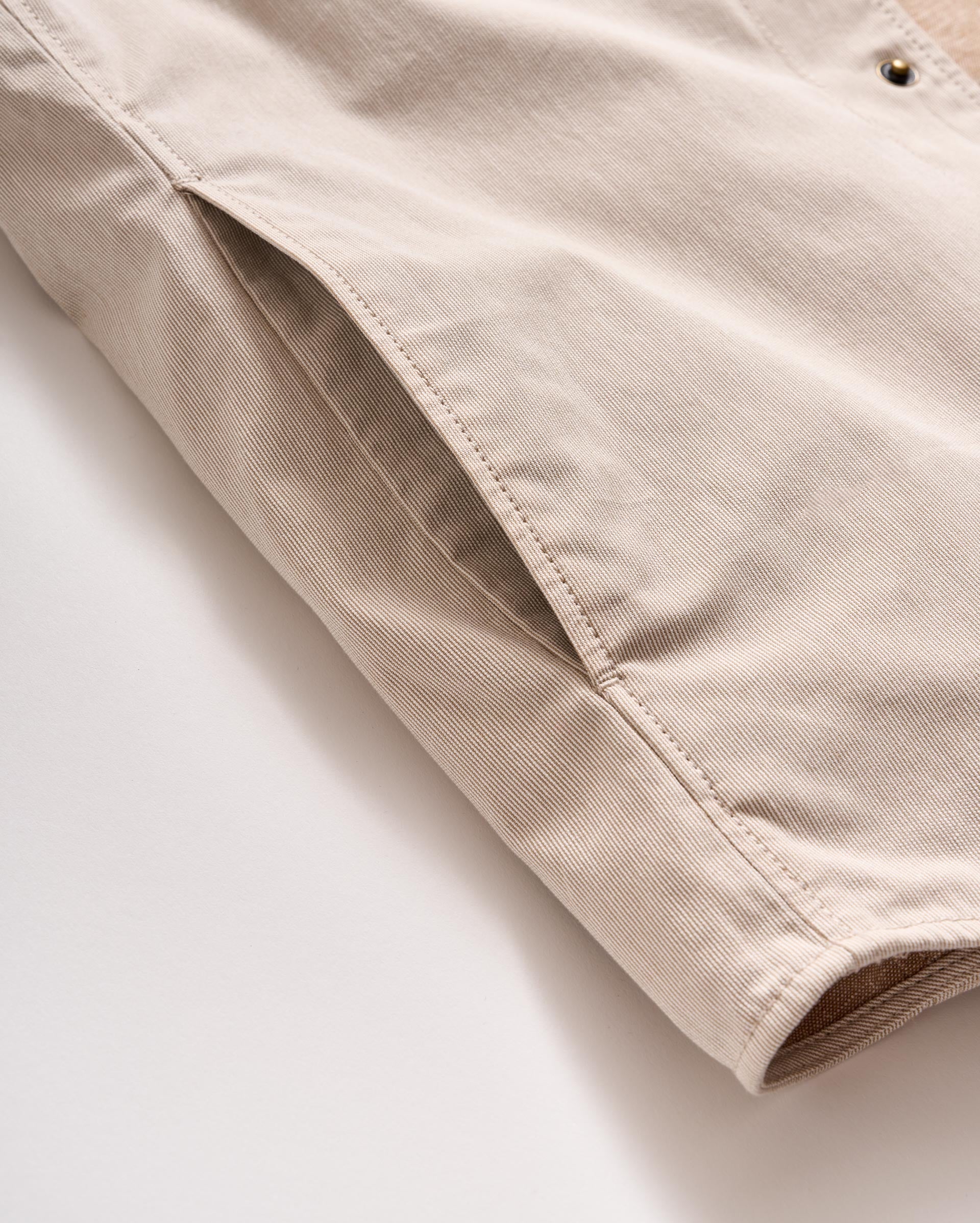A close-up of a beige fabric jacket, focusing on a side pocket and part of the sleeve, with visible stitching and metal snap button detail.