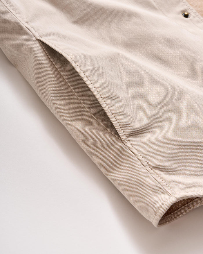 A close-up of a beige fabric jacket, focusing on a side pocket and part of the sleeve, with visible stitching and metal snap button detail.