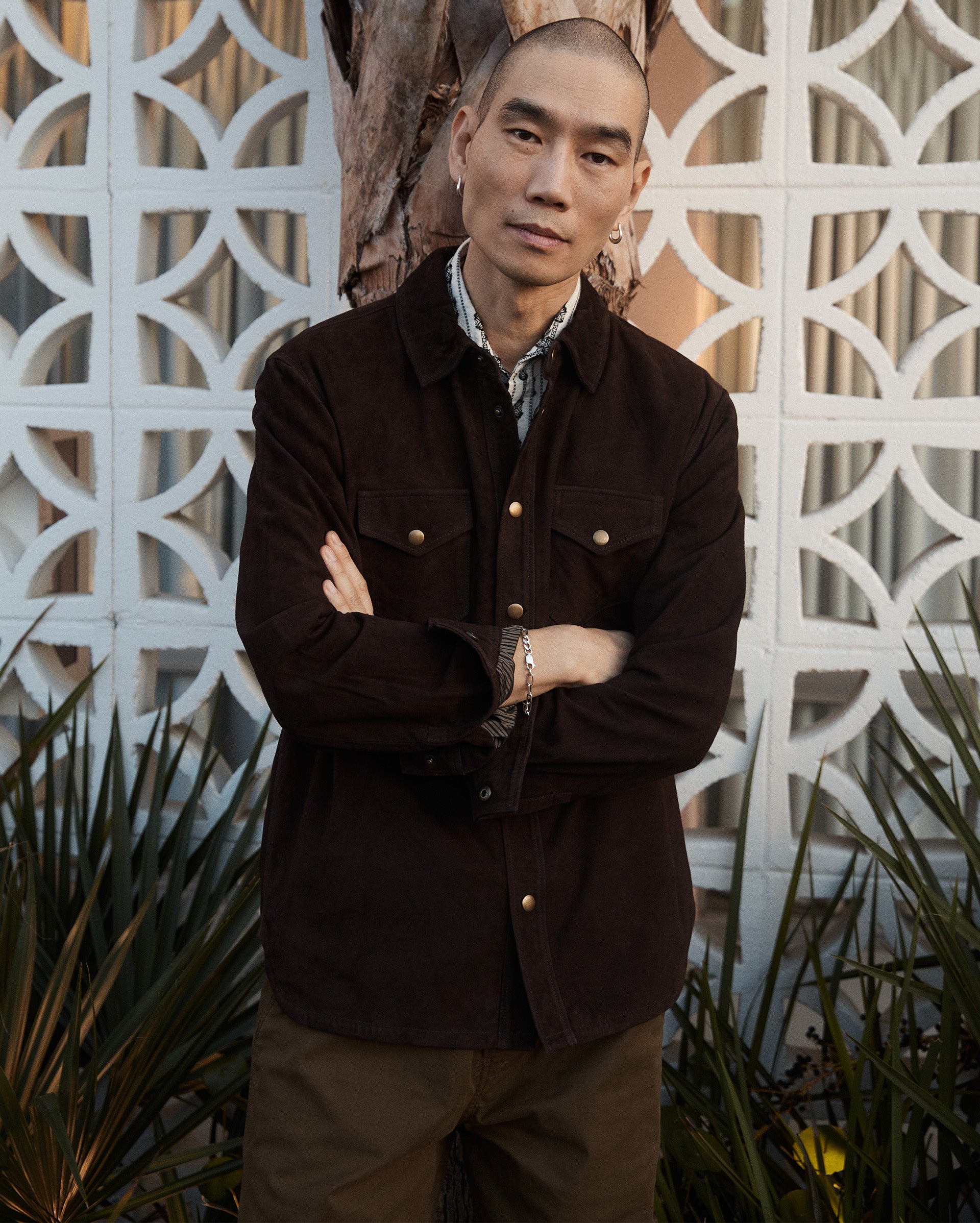 A person with a shaved head stands outdoors in front of a patterned white wall and a tree, wearing a dark brown buttoned jacket over a collared shirt, with arms folded and a neutral expression. Shrubs are visible at the bottom.