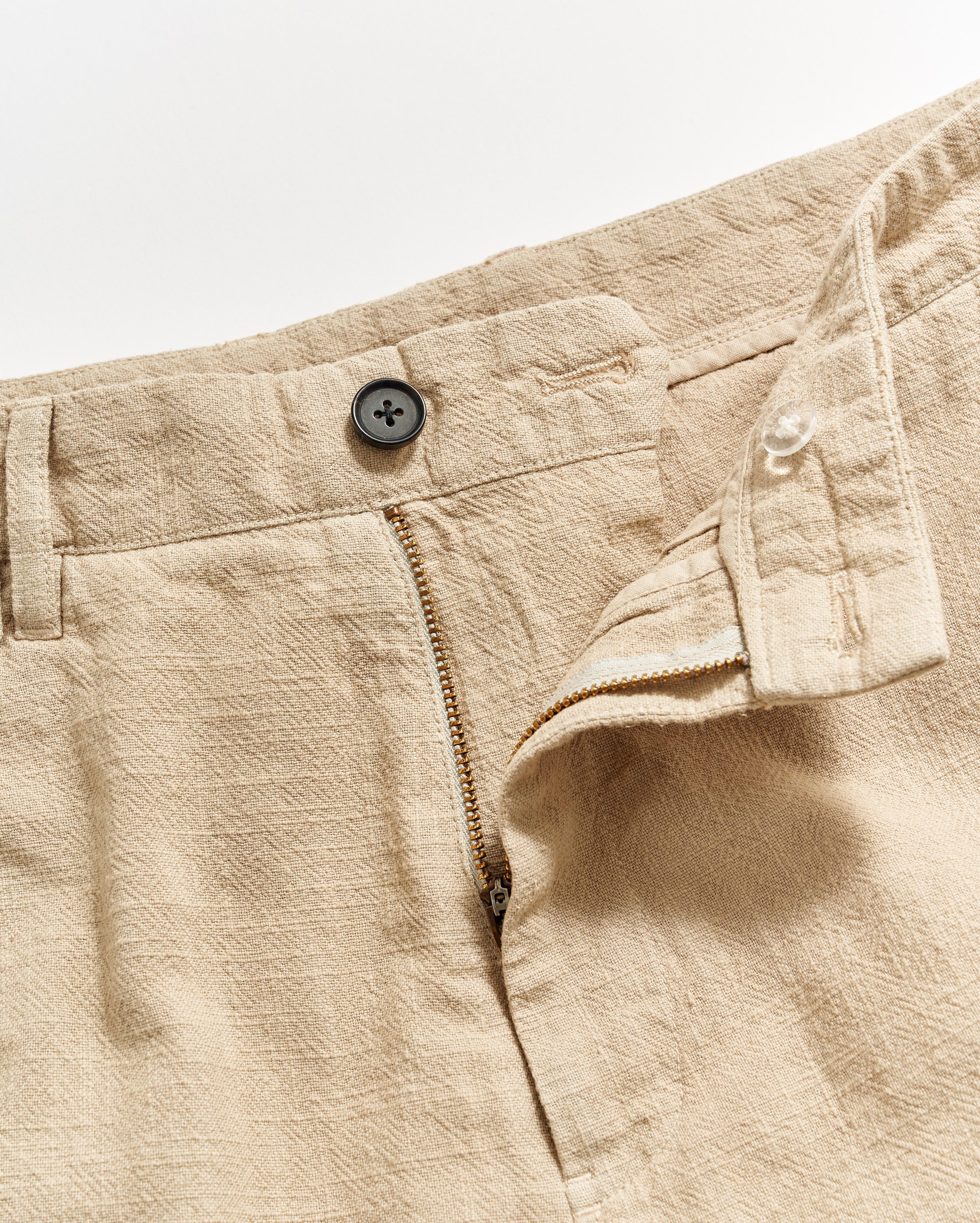 Beige textured fabric pants with a partially open zipper fly, a black button at the waistband, and an inner button closure, shown on a white background.
