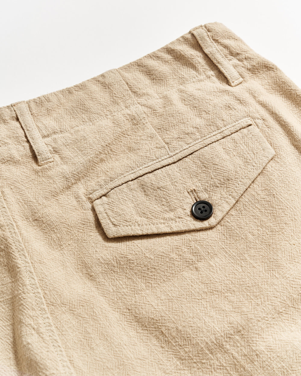 Close-up of beige textured pants showing a back pocket with a flap and a black button, along with belt loops and visible stitching details.