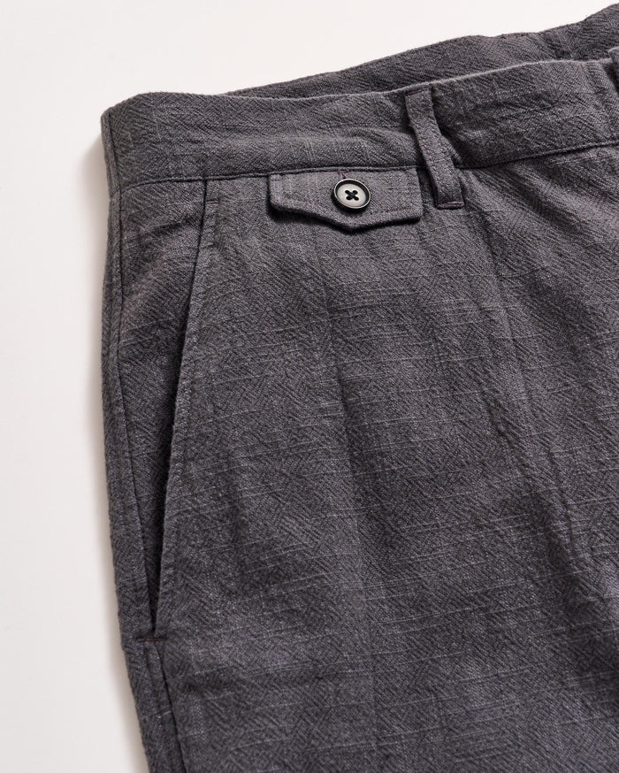Close-up of the upper section of a pair of textured gray pants, featuring a waistband with a belt loop, a small decorative flap with a black button, and a pleated design near the pocket.