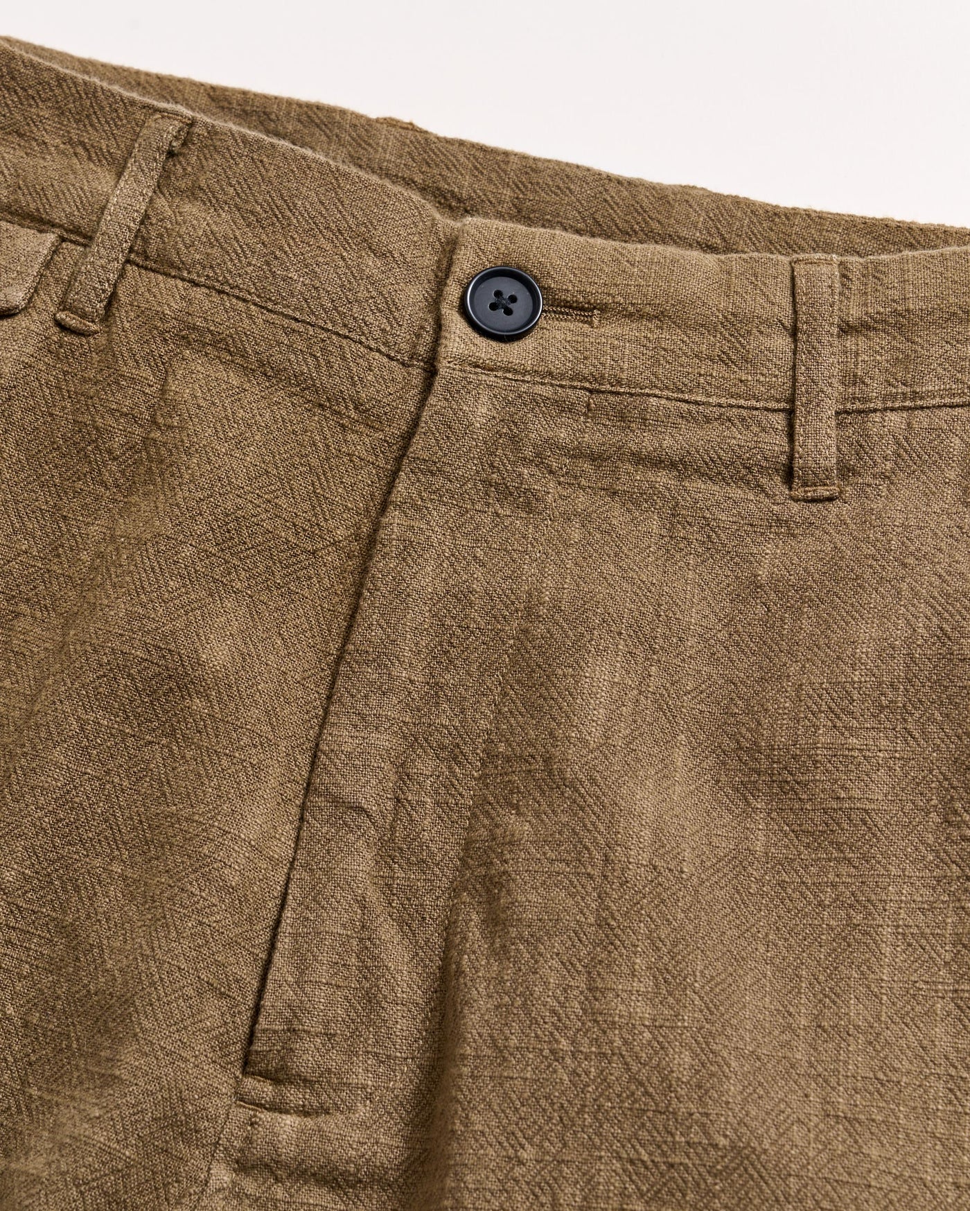 Close-up of the waistband and front of a pair of brown textured pants with a black button, belt loops, and a visible seam on a light background.