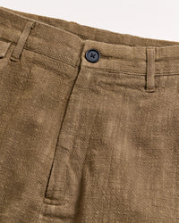 Close-up of the waistband and front of a pair of brown textured pants with a black button, belt loops, and a visible seam on a light background.