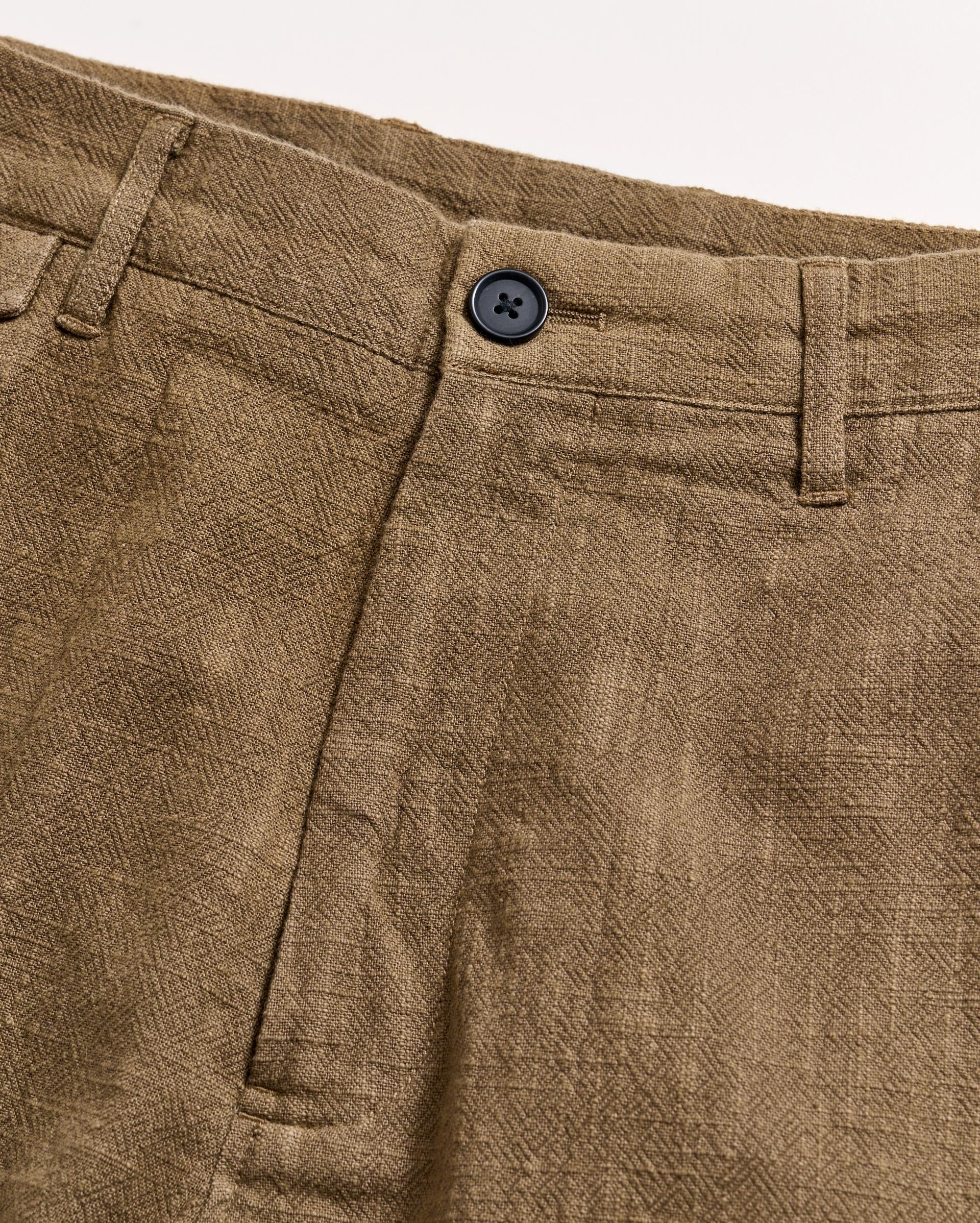 Close-up of the waistband and front of a pair of brown textured pants with a black button, belt loops, and a visible seam on a light background.
