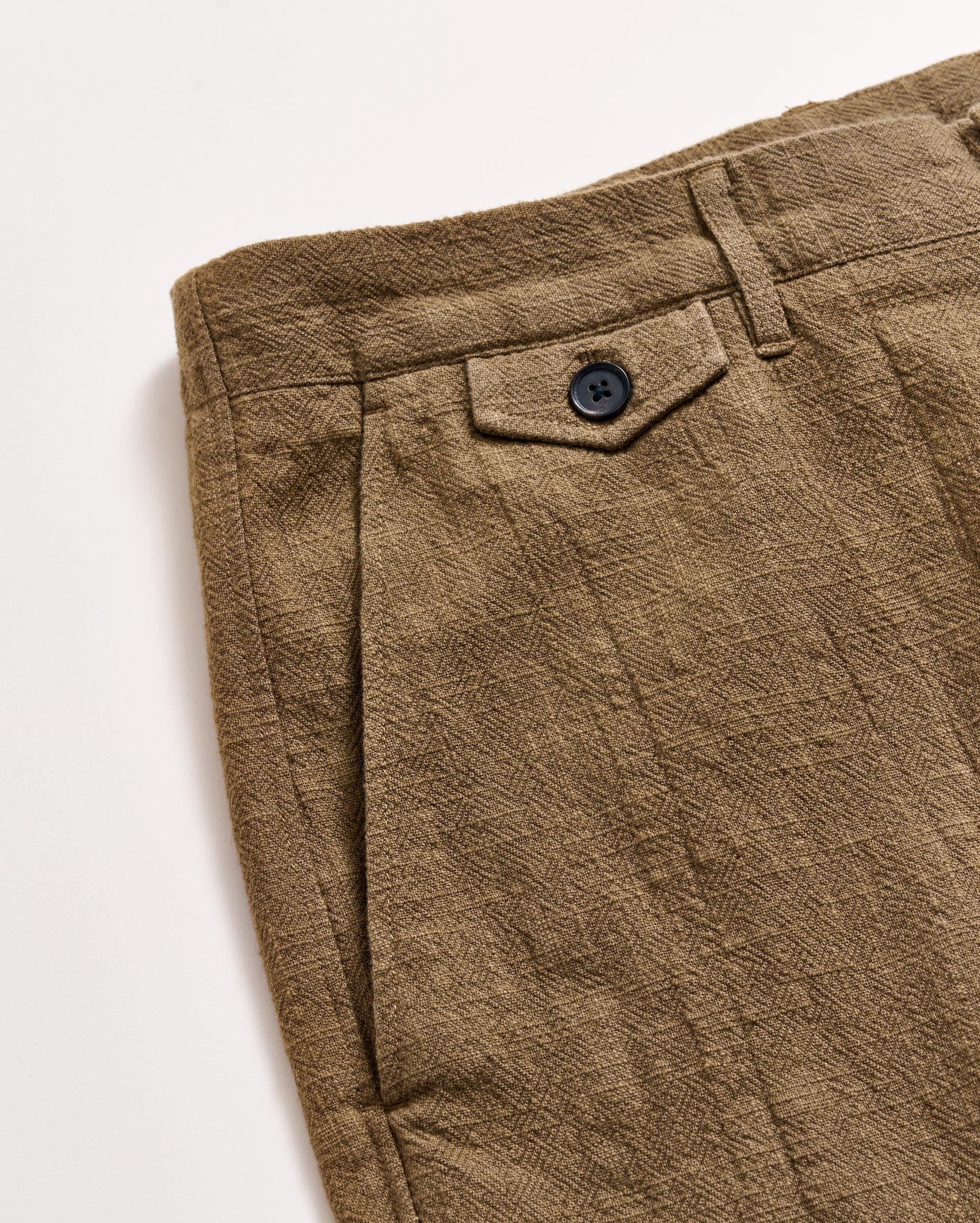 Close-up of a pair of brown textured pants featuring a front pleat, a waistband with a belt loop, and a small flap pocket secured with a black button. The fabric has a crinkled, patterned finish.