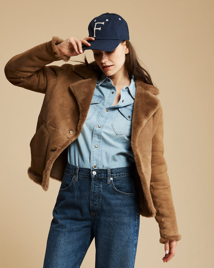 Woman wearing a brown jacket, blue denim shirt, and jeans with a navy cap on a beige background