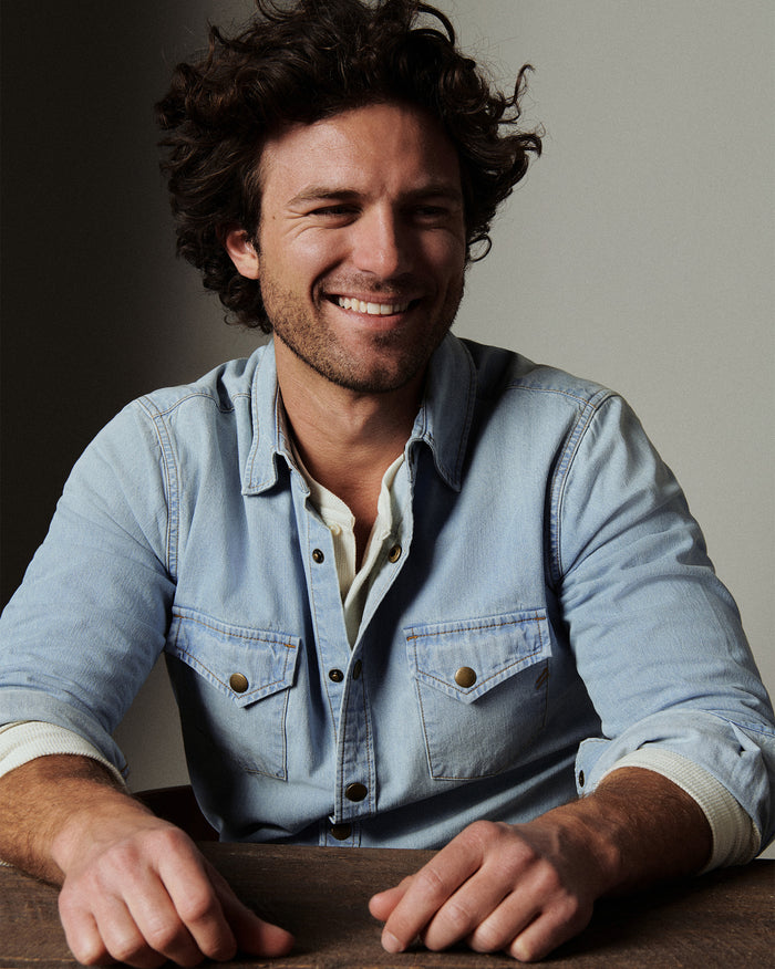 Man wearing a light blue denim shirt sitting at a table with a neutral background