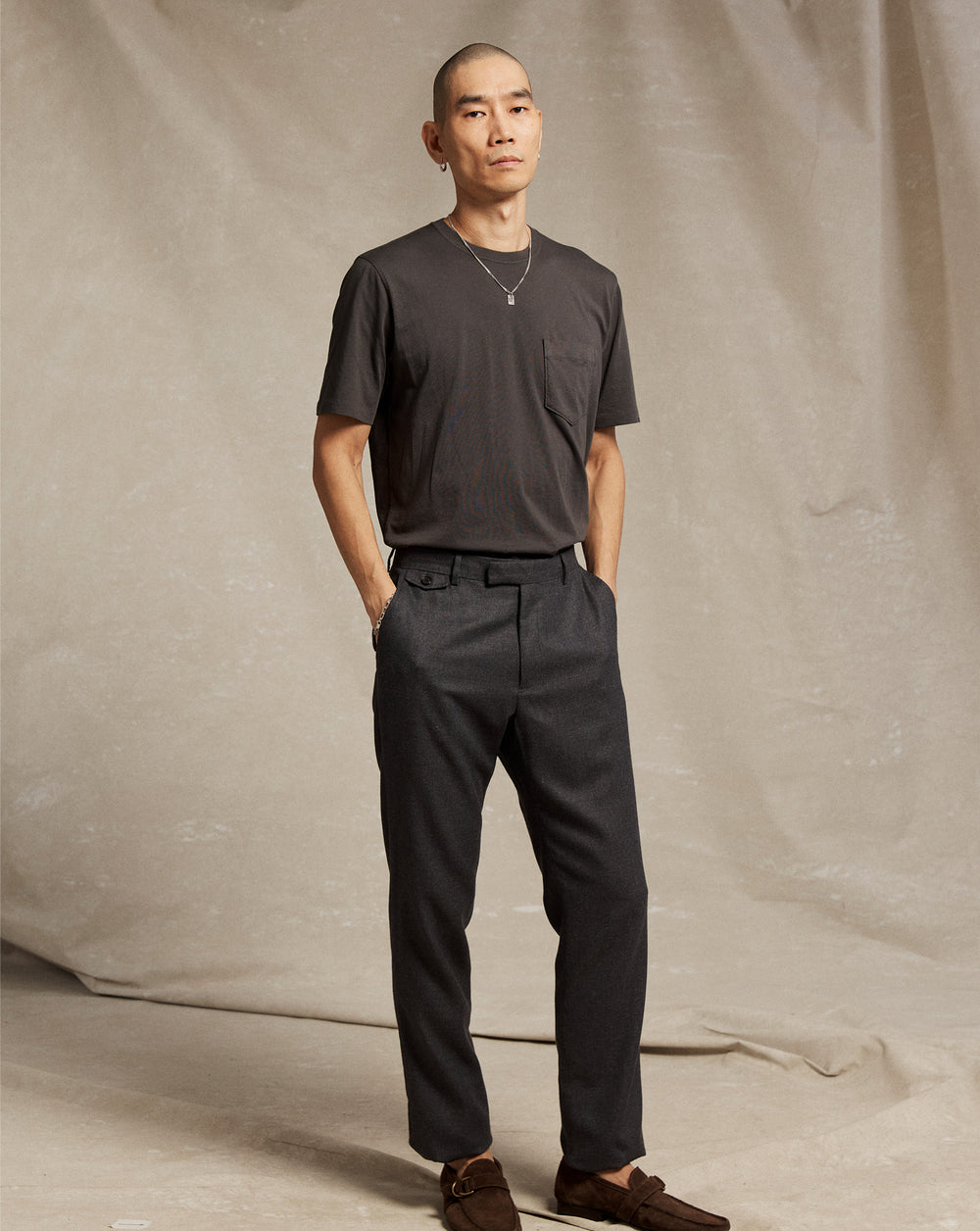 A person with a shaved head stands against a beige fabric backdrop, wearing a dark short-sleeve t-shirt, dark tailored pants, brown shoes, and a necklace, with hands in their pockets and a neutral expression.