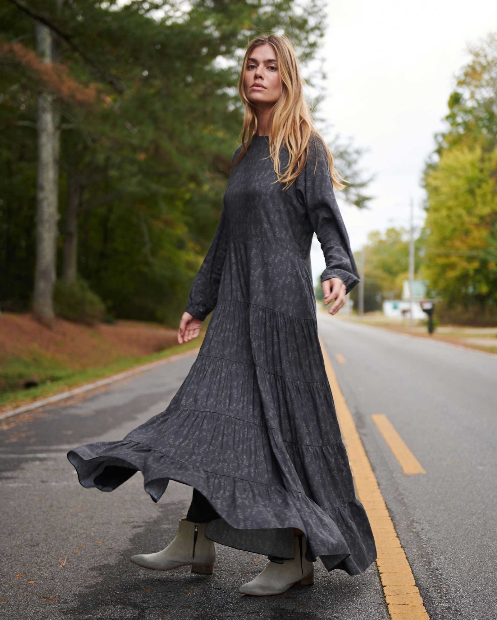 A woman with long blond hair wears a flowing, dark gray dress and light-colored boots, walking gracefully on an empty road lined with trees on a cloudy day.