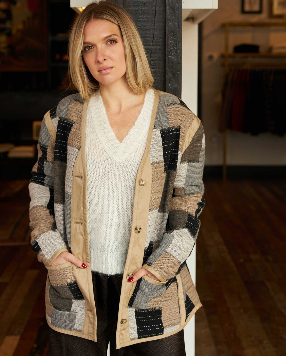 A woman with shoulder-length blonde hair stands indoors, wearing a white V-neck sweater and a patchwork-patterned cardigan with neutral colors. She has her hands in her cardigan pockets and looks calmly at the camera.