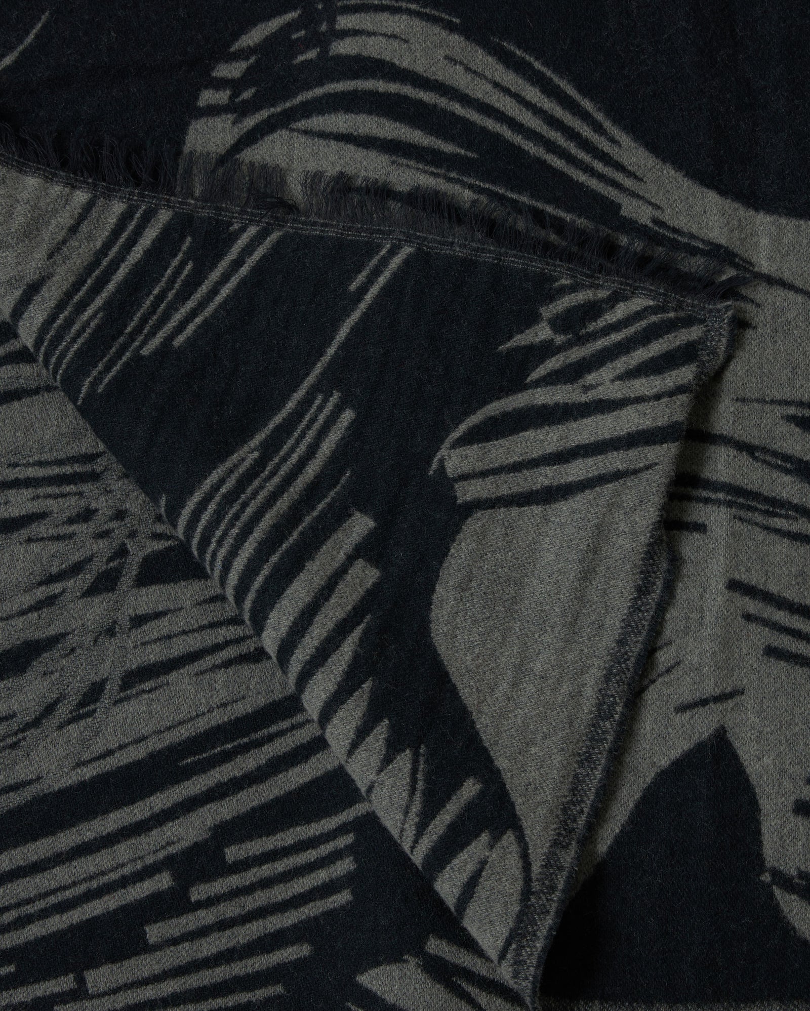 A close-up of a textured fabric with abstract black and gray patterns and fringed edges, showing a folded corner.